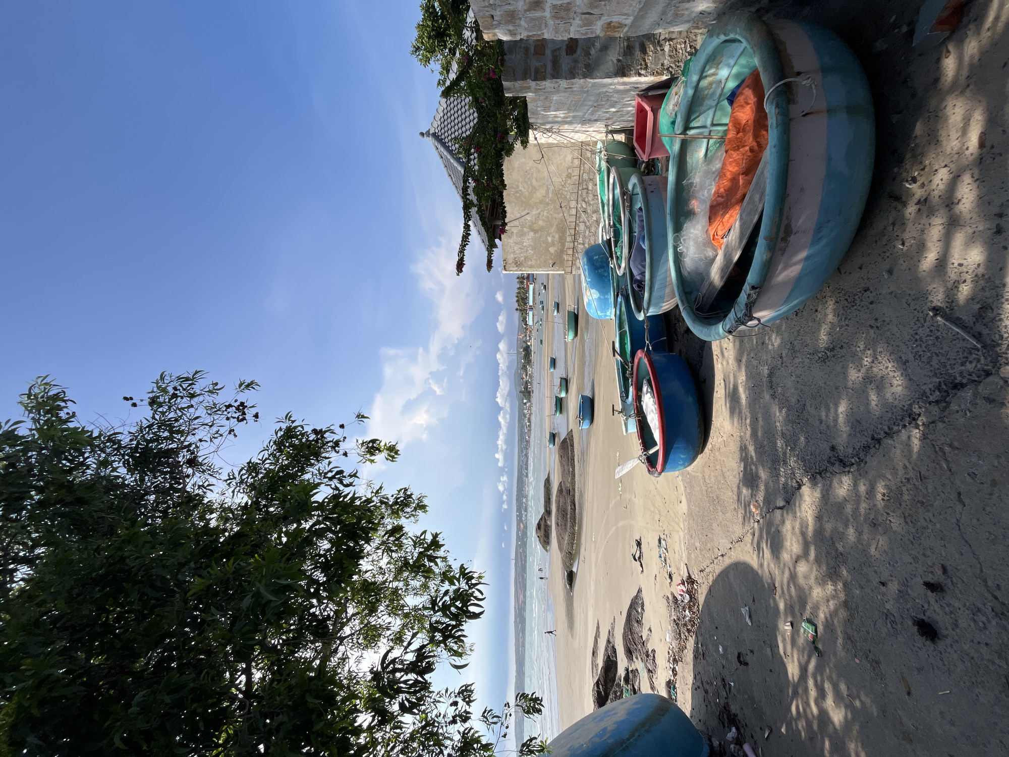The image depicts a coastal scene in Mũi Né, Vietnam, with a focus on a collection of round, basket-like fishing boats beached on a sandy shore. The boats, predominantly in shades of blue and green, are clustered on the sand, with some containing fishing nets and orange tarps. In the foreground, a path made of concrete and sand leads towards the beach, littered with small pieces of debris and casting shadows from overhead foliage.
Further down the beach, several more boats are visible, as well as a few scattered rocks. The sea stretches out to the horizon, with gentle waves and a distant shoreline dotted with buildings. The sky is clear blue with scattered white clouds, indicating fair weather. To the right, a stone wall and a building with a tiled roof are visible, suggesting the edge of a settlement or a property bordering the beach. There are a few distant figures walking on the beach and in the water. The overall impression is of a tranquil, working fishing village on a sunny day.