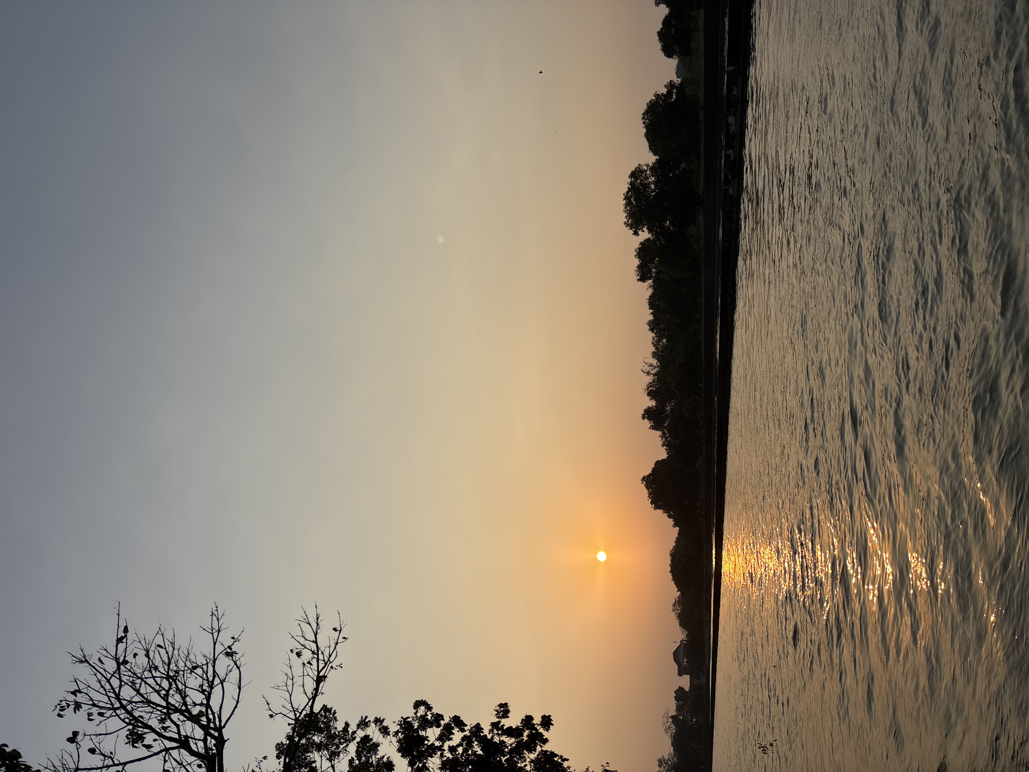 The image depicts a serene sunset over a body of water, likely a river or lake, in Beber, Indonesia. The sun, a bright orange orb, is low on the horizon, casting a warm glow across the sky and reflecting on the rippling water. Silhouetted trees line the opposite bank, with a small building visible among them. The foreground is dominated by the water, its surface disturbed by gentle waves that catch the sunlight. A bare tree branch extends into the frame from the upper left corner, its dark silhouette contrasting with the pale, gradient sky. The overall atmosphere is tranquil and natural, characteristic of a rural or semi-rural Indonesian landscape at dusk.
