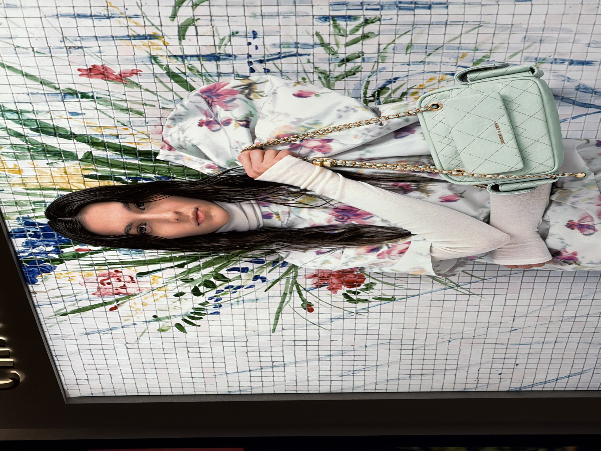 A young woman with dark, wet hair stands in front of a wall with a mosaic of floral paintings. She is wearing a white turtleneck and a white blouse with a floral pattern. She is holding a light green quilted handbag with a gold chain strap. The handbag has a quilted pattern and a small pocket with the "CHARLES & KEITH" logo. The background mural depicts a loose, watercolor-style floral arrangement with green, blue, red, and yellow hues. The woman's expression is neutral, and she looks directly at the camera. The scene appears to be set in a well-lit retail space, possibly a boutique or a department store, given the presentation of the handbag. The location is Kuta, Indonesia.