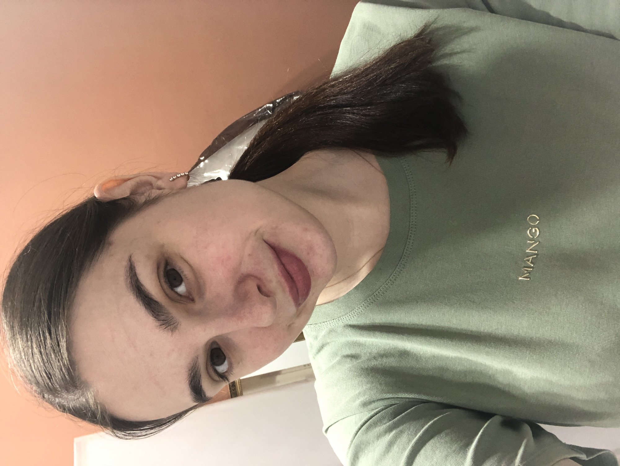 A woman with dark brown hair, parted on the left and pulled back, is facing the camera. She has fair skin, brown eyes, and is wearing light-colored lipstick. In her left ear, she has a small earring with multiple stones. Her left side of her hair has foil on it suggesting that she is currently having her hair dyed. She is wearing a light green T-shirt with the word "MANGO" printed in gold on the right chest area. The background is a two-toned wall, with the top half appearing to be a warm brown or peach color. The location in Chaikovsky, Russia is not possible to confirm from the image.
