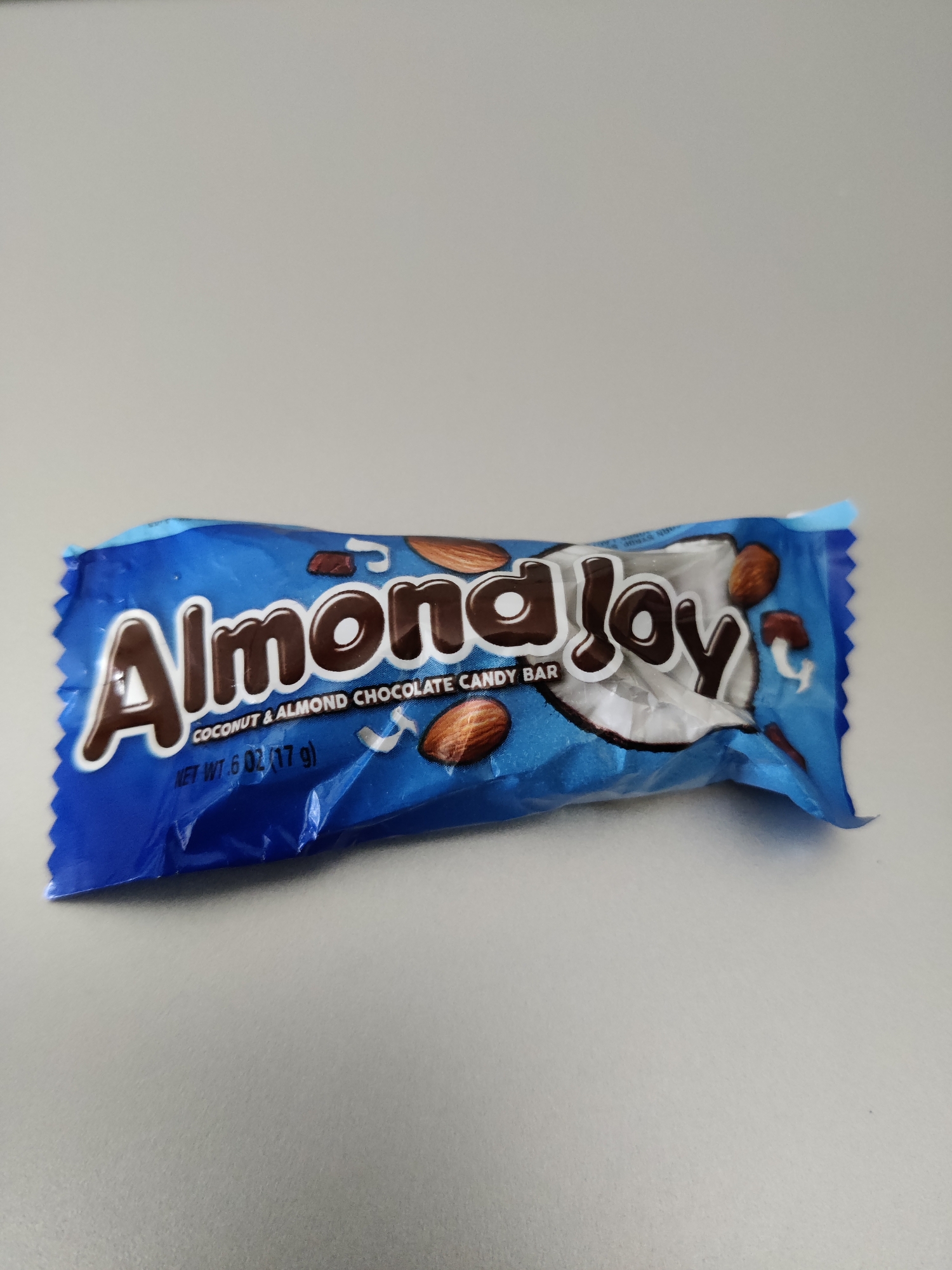 A blue wrapper for an Almond Joy candy bar is laid out horizontally on a plain, light gray surface. The wrapper is partially crumpled. The brand name "Almond Joy" is prominently displayed in large, dark brown letters with a white outline. Below the brand name, it reads "COCONUT & ALMOND CHOCOLATE CANDY BAR" in smaller white text, and further down, "NET WT. 6 OZ (17 g)" also in white text. Images of almonds and coconut pieces are visible on the wrapper. The background is an out-of-focus, uniform light gray. There are no people or other objects in the scene. The location is Lower Paxton Township, United States of America.