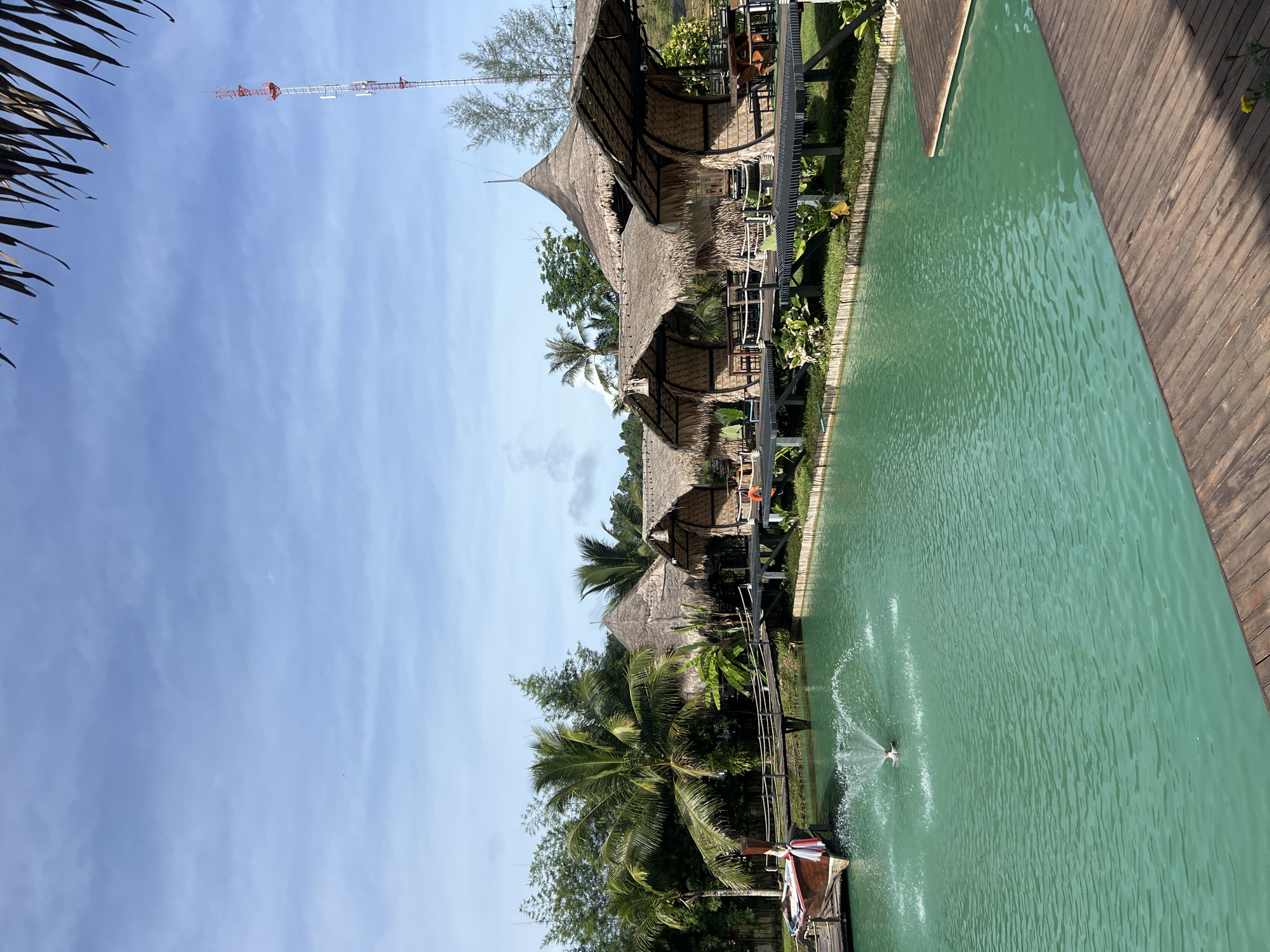 The image depicts a serene outdoor scene at Tambon Pa Kae Bo Hin, Thailand. A tranquil, jade-colored body of water, possibly a swimming pool or a calm bay, dominates the right side of the frame. Wooden decking lines the edge of the water, with the sun casting shadows across its planks.
Along the left side of the water, a series of traditional thatched-roof structures are visible, nestled amongst lush tropical foliage. These buildings, perhaps bungalows or a restaurant, feature open-air seating areas with tables and chairs, suggesting a place of relaxation or dining. Palm trees and other dense greenery surround the structures, adding to the tropical ambiance.
A small fountain sprays water in the middle of the body of water, creating gentle ripples. In the background, a tall communication tower rises against a clear blue sky with scattered white clouds. The overall impression is one of a peaceful, sunny day in a tropical resort or retreat. The clear sky suggests it is daytime, and the quality of light implies it is not the hottest part of the day. No individuals are clearly discernible in the image, nor is any text visible.