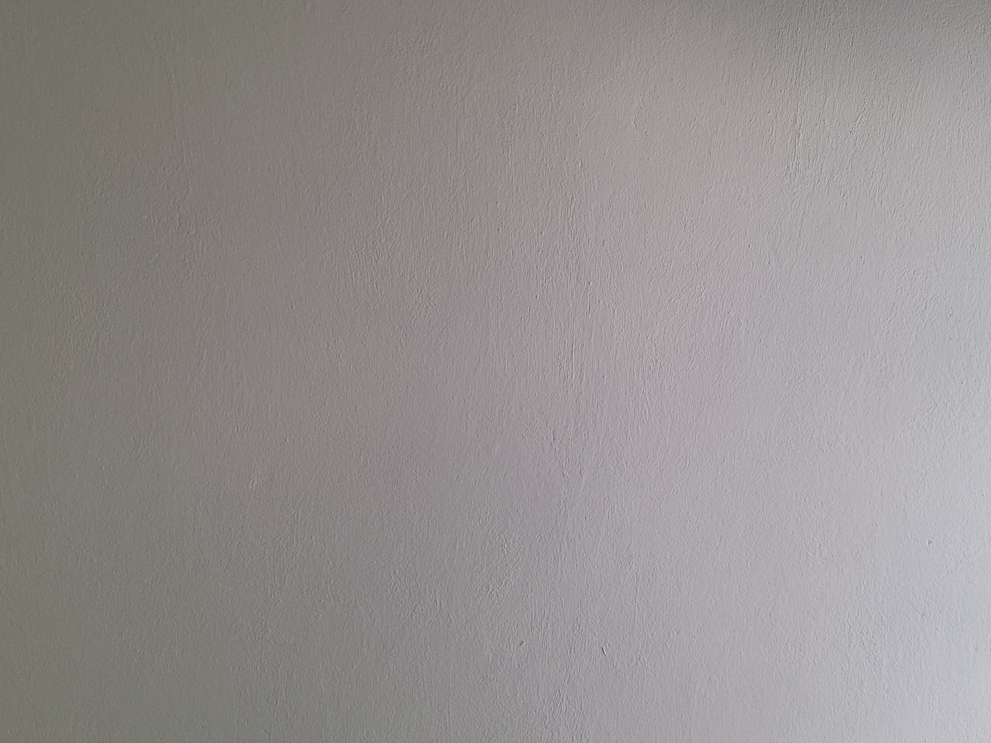 The image is an abstract, close-up shot of a textured, light gray wall. The texture appears to be from plaster or stucco, with subtle undulations and slight imperfections visible across the surface. The lighting creates a soft gradient, with the top of the image appearing slightly darker and the bottom lighter, suggesting the light source is from below or to the side. There are no people, objects, or discernible features that would indicate a specific location within Varna, Bulgaria, or any other city. The image solely depicts the texture and color of a wall.