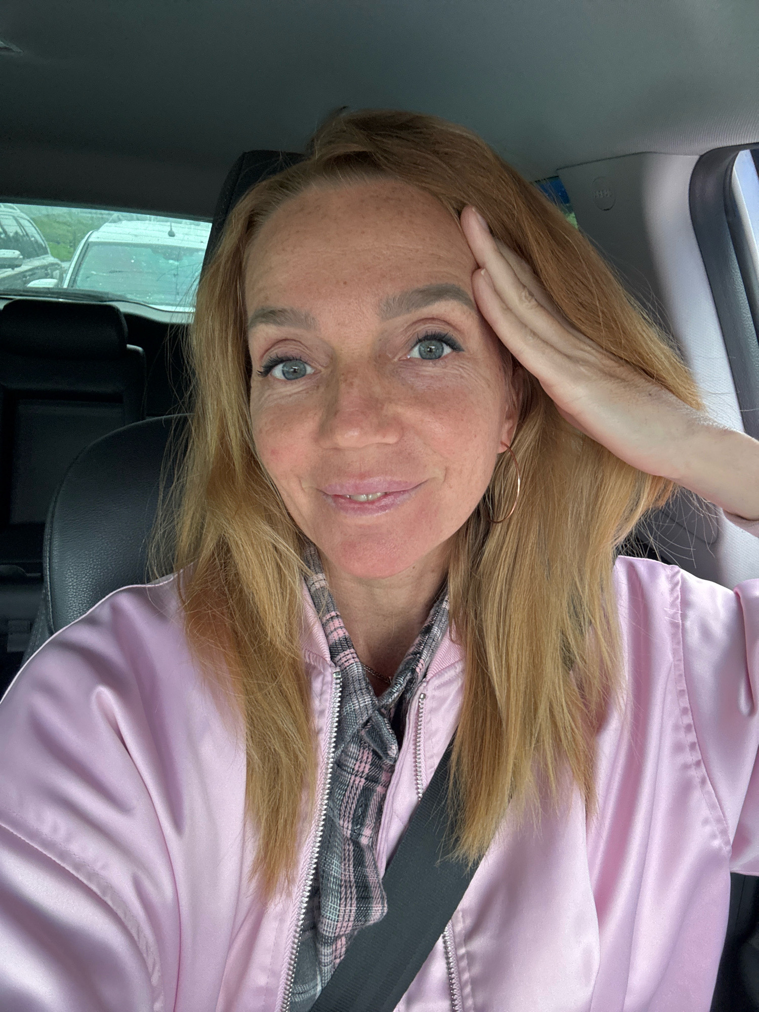 A fair-skinned woman with blonde hair and blue eyes, wearing a pink satin jacket and a grey and pink plaid shirt, poses for a selfie in a car. Her right hand is raised to her forehead, and she is smiling. In the background, the interior of the car is visible, including a black leather seat and a glimpse of vehicles outside through the rear window. The environment suggests an urban setting, potentially within the city of Saint Petersburg, Russia, given the context. The lighting is overcast, indicating an outdoor environment under a cloudy sky.