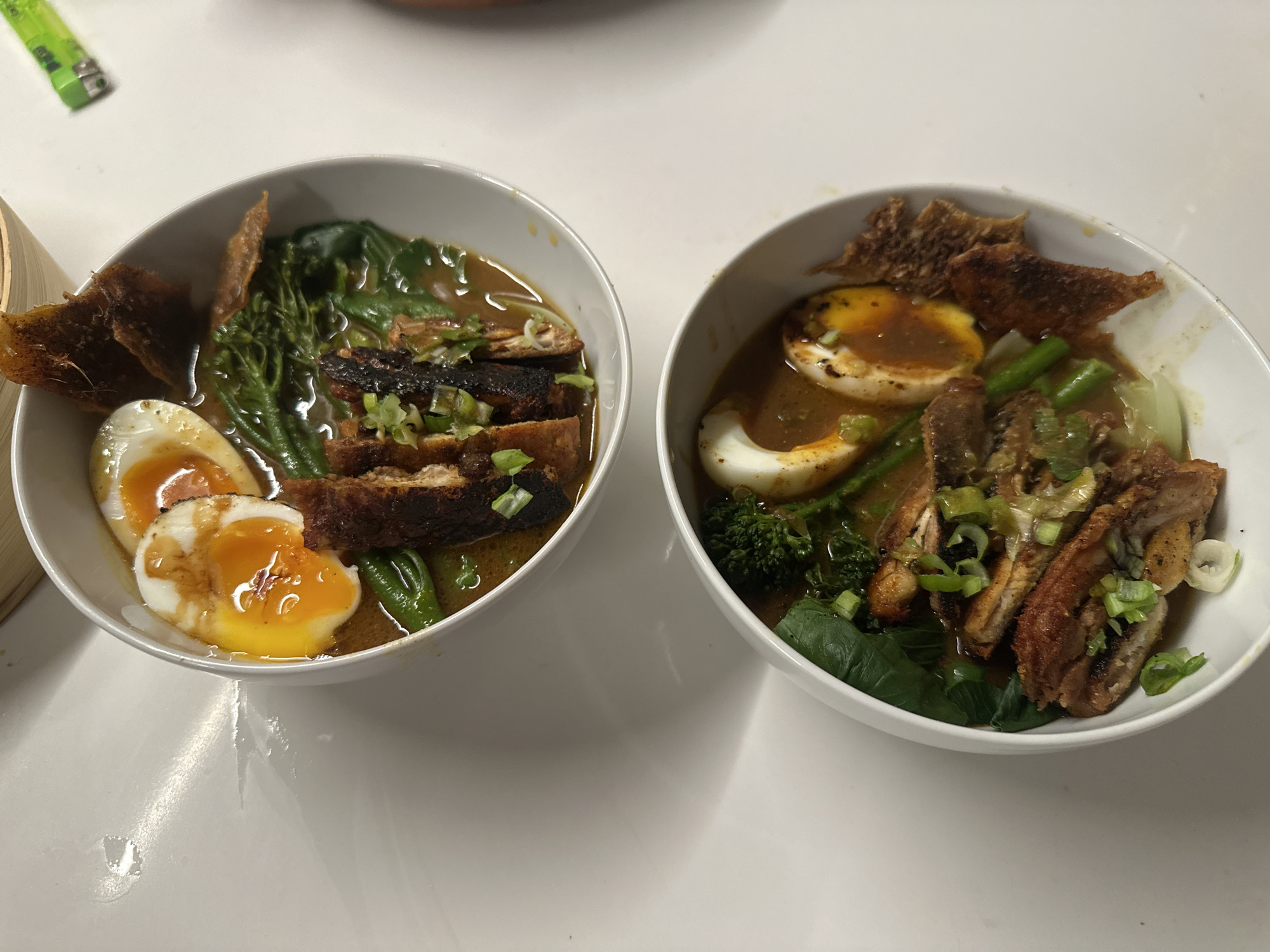 Two bowls of ramen are arranged on a white surface. Each bowl contains broth, sliced eggs, greens, and what appears to be sliced meat. The eggs are soft-boiled with runny yolks. The ramen on the left includes green beans, while the ramen on the right includes broccoli florets. The scene is indoors. There is no indication of location in London. No people are visible.