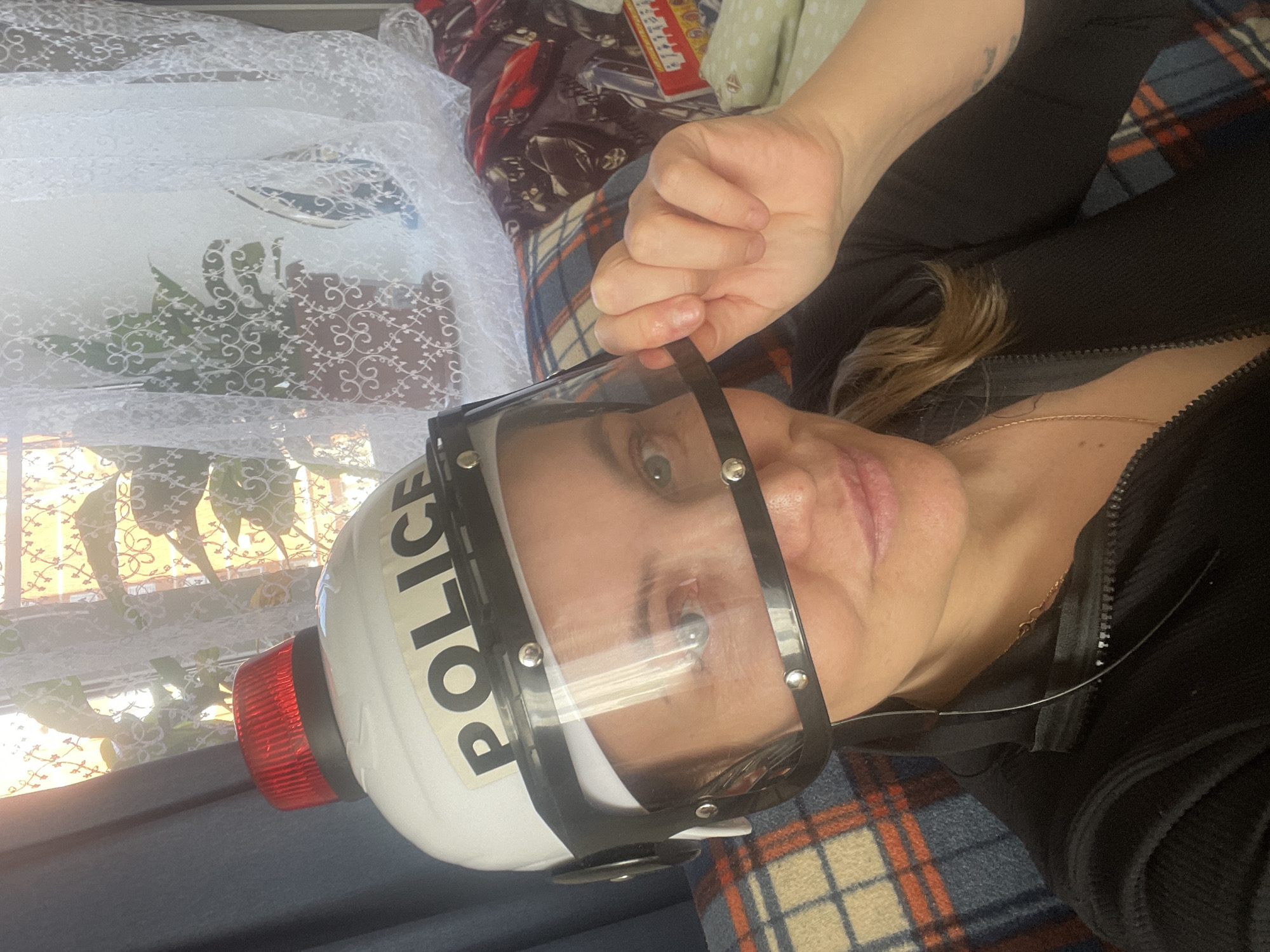 A woman is lying on a bed wearing a white police helmet with the word "POLICE" printed on the side. The helmet has a clear face shield attached and a red flashing light on top. She is wearing a black zippered top and a gold necklace. The bed has a patterned comforter and a white lace curtain is visible in the background. Based on the presence of curtains, and other soft goods, it can be inferred that the location is most likely indoors. The location cannot be accurately pinpointed to Chaikovsky, Russia based solely on the contents of the image.