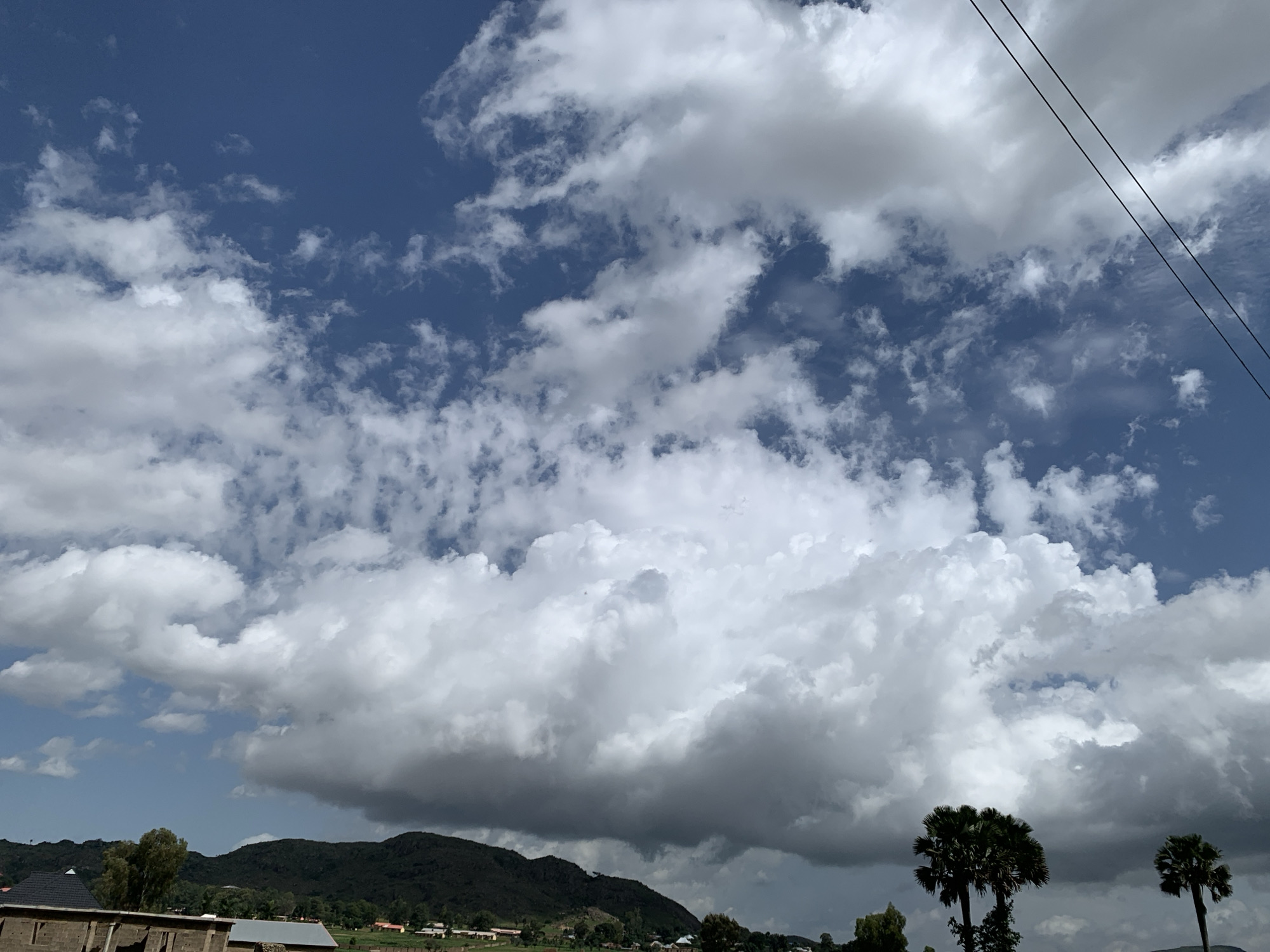 The image depicts a landscape under a dynamic sky. In the foreground, the upper portion of a brick structure is visible, suggesting construction or a building in progress. Beyond this, a line of dark green hills forms the midground, dotted with scattered trees and some small buildings indicating a rural or semi-rural settlement. The sky dominates the upper two-thirds of the frame, filled with a dramatic display of cumulus clouds. Some clouds are bright white, catching the sunlight, while others are darker, casting shadows and suggesting an approaching weather system or recent rain. Two palm trees stand prominently in the right midground. In the far upper right corner, two parallel black wires, likely power lines, stretch diagonally across the frame, indicating the presence of infrastructure. The scene is rendered in natural light, with the bright clouds and blue sky suggesting a daytime setting. The location appears to be Kuru Karama, Nigeria.