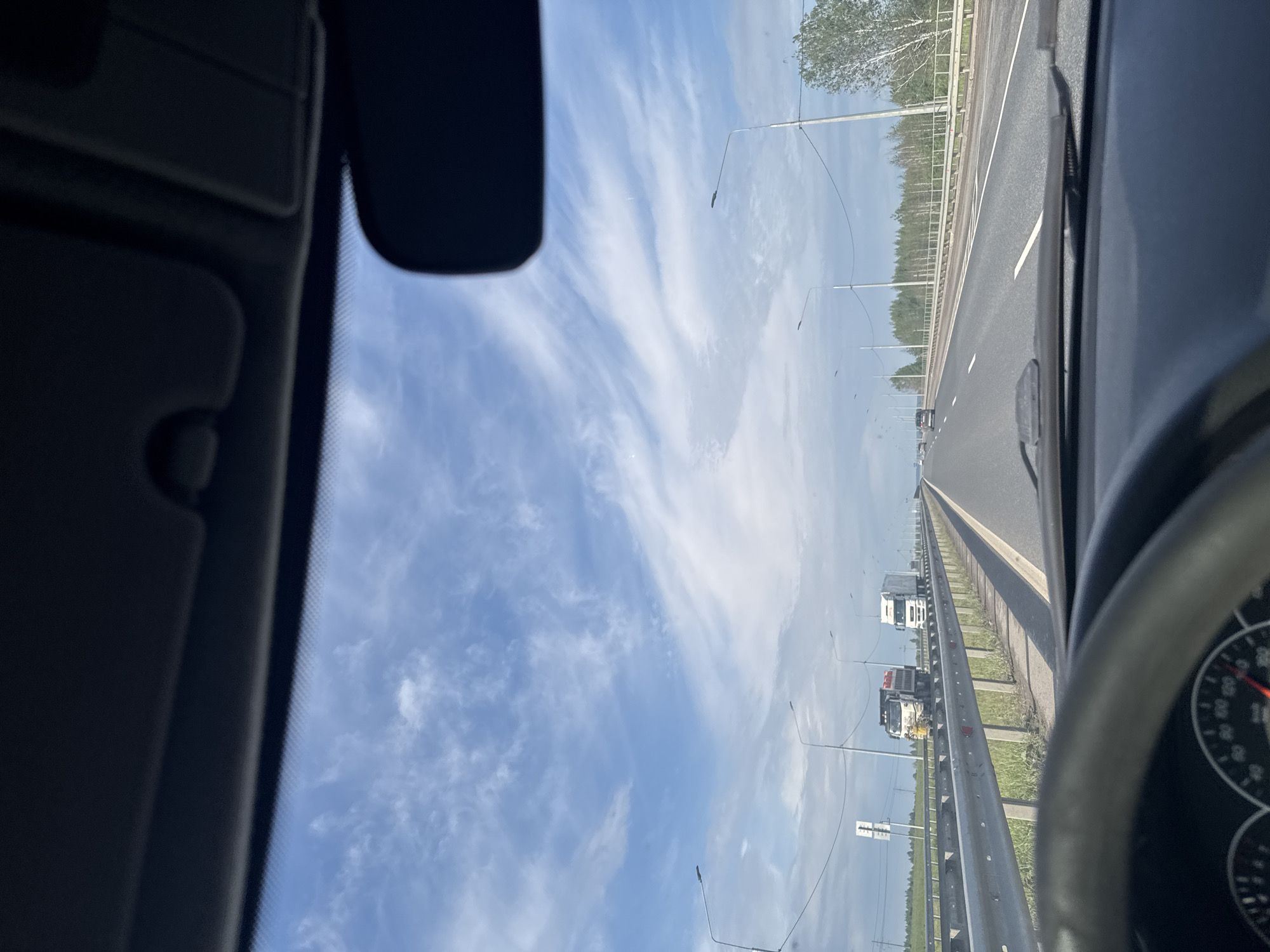 The image displays a daytime view from the interior of a vehicle, looking forward onto a multi-lane highway. Elements of the car's interior frame the scene, with the roof lining and sun visor visible in the top-left and a partial view of the dashboard and steering wheel on the right.
The highway stretches into the distance under a bright blue sky, which features scattered, wispy white clouds, indicating clear and fair weather. The road has distinct white lane markings and is bordered on the right by a metal guardrail. Beyond the guardrail, a green verge leads to a dense line of trees, suggesting a natural or rural environment.
Street lights or utility poles line the side of the highway, receding into the background. Several large trucks are visible in the distance, traveling on the road. The scene captures a moment of highway travel, consistent with a location such as Усть-Сыны, Russia. No discernible text is visible within the image.