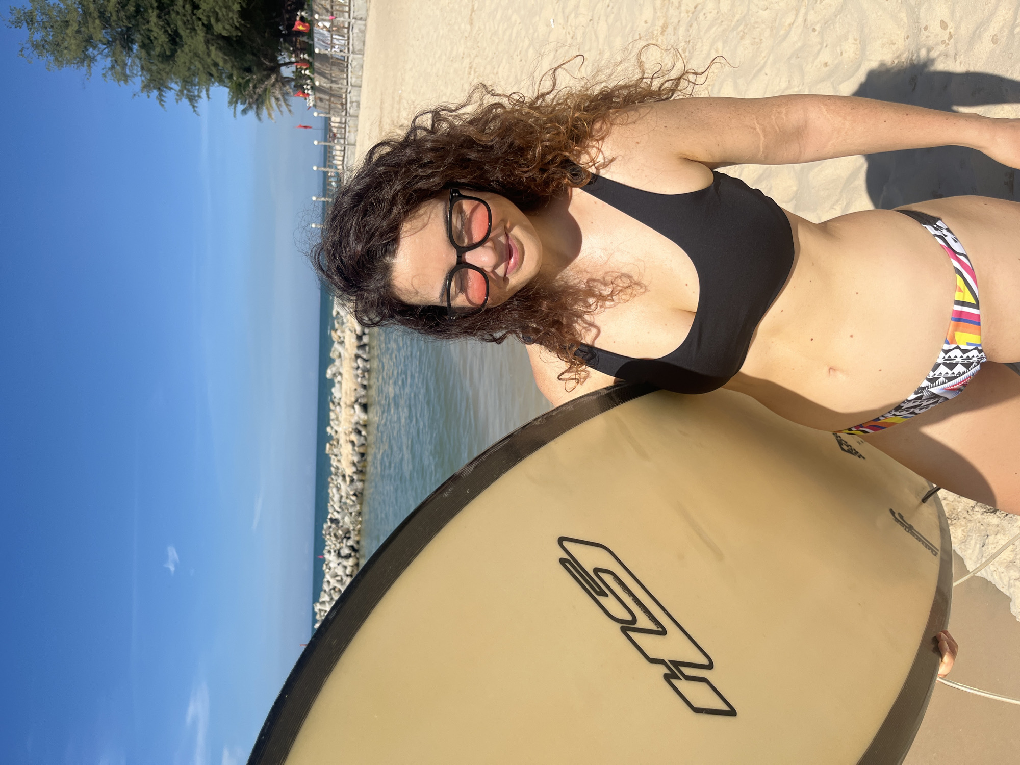 A woman stands on a sandy beach in Phu Thuy Ward, Vietnam, holding a surfboard. She is wearing a black bikini top and a colorful bikini bottom. She has curly brown hair and is wearing pink-tinted sunglasses. The ocean is visible behind her, with a breakwater made of large rocks in the distance. The sky is clear and blue, with a few wispy clouds. Trees and a wooden structure with flags are visible on the right side of the image. The surfboard is light beige with black lettering.