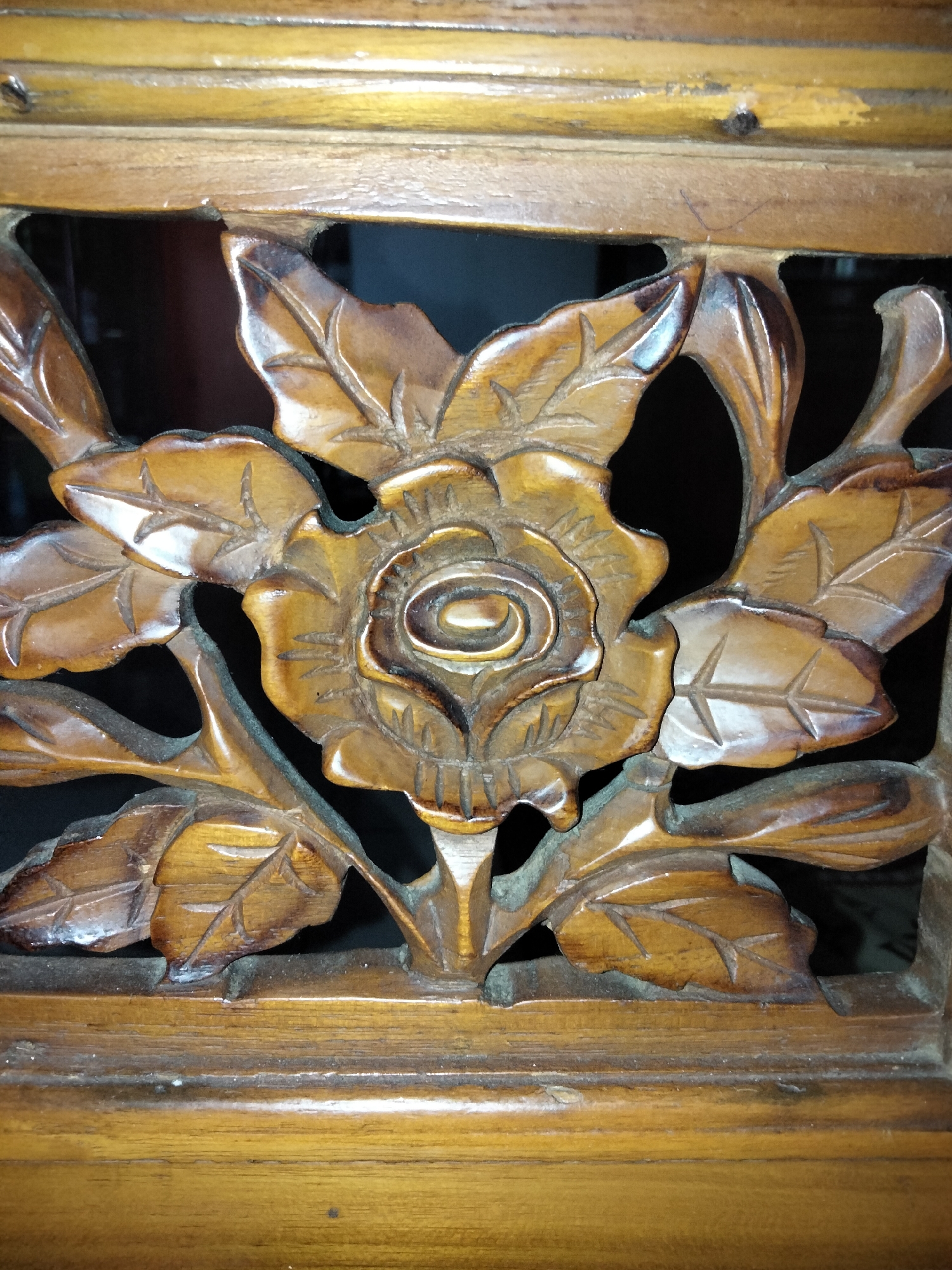 The image shows a close-up of a wooden carving of a rose with leaves. The carving is intricate and detailed, with the petals of the rose and the veins of the leaves clearly visible. The wood is a warm, rich brown color, and it appears to be well-maintained. The carving is set within a wooden frame, and the background is dark, which makes the carving stand out. The lighting is focused on the carving, highlighting its texture and form. The image does not provide any information about the location in the city of Payagaboh, Indonesia.