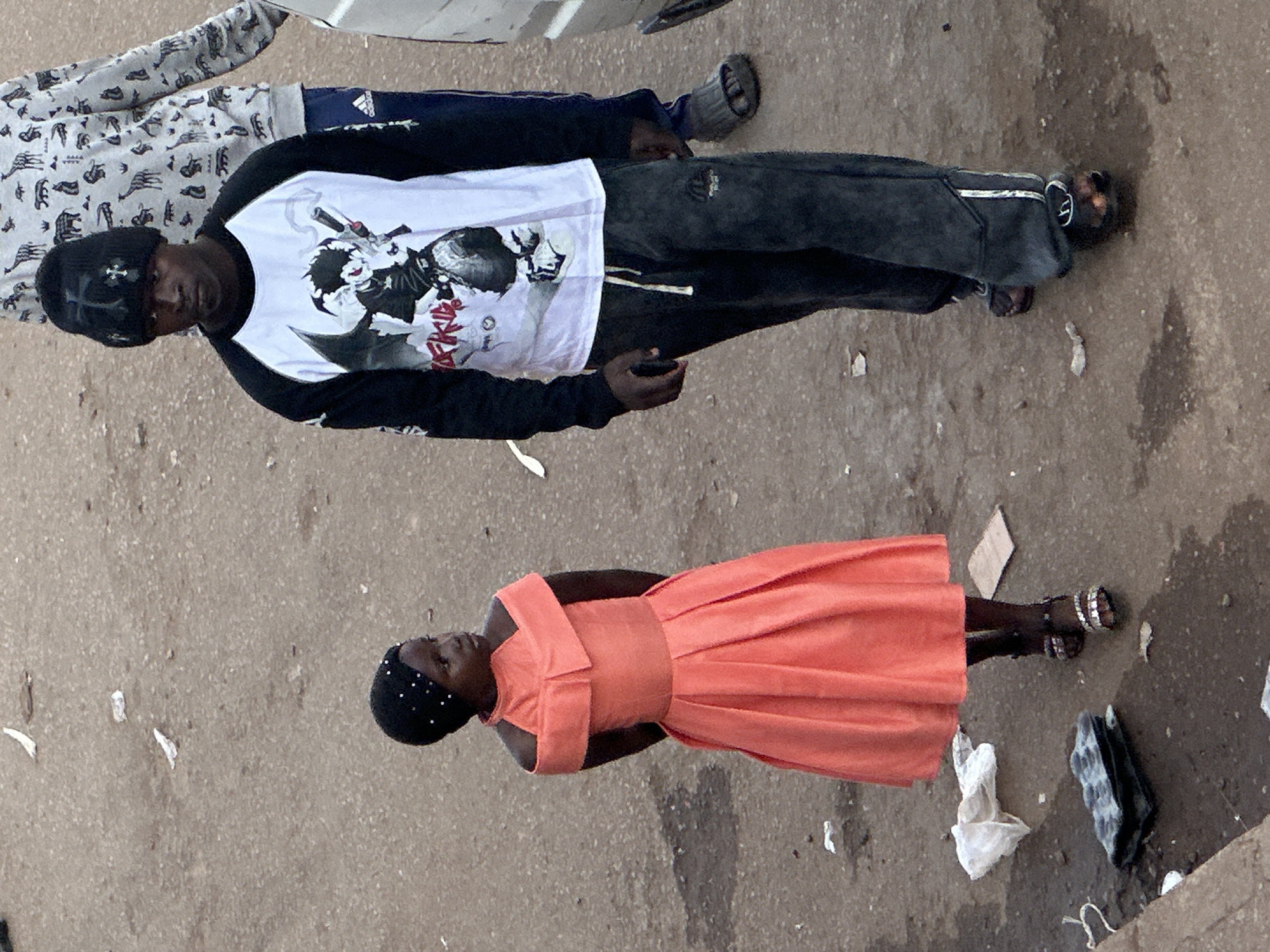 In Bukuru, Nigeria, an outdoor scene unfolds on a dusty, dirt road littered with white paper scraps and debris. A young girl, adorned in a bright orange off-the-shoulder dress with gathered fabric and a full skirt, stands looking to her right. She wears a black beaded headscarf. Beside her, a young man wearing a black beanie with cross embellishments, a black and white raglan-sleeved shirt featuring an anime-style graphic, and dark gray sweatpants walks towards the viewer. He is holding a dark object in his right hand, possibly a phone, and wears dark sandals. In the background, another person is partially visible, wearing a gray hooded sweatshirt with a black and white animal print pattern and dark blue pants with a white stripe.