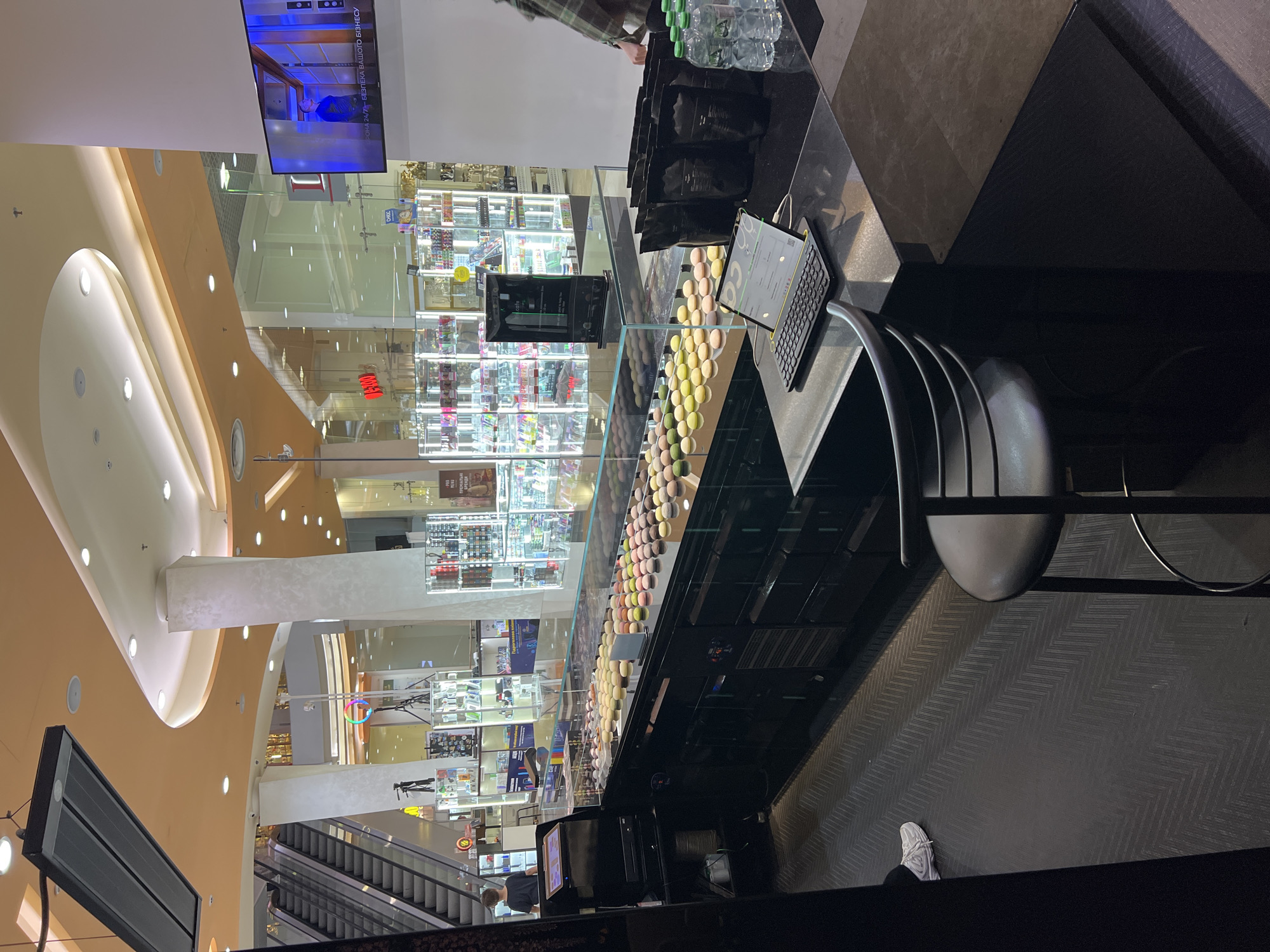 The image depicts a brightly lit interior of a commercial establishment, likely a shop or café, possibly located in Odesa, Ukraine. The foreground features a display counter filled with a colorful assortment of macarons. Behind the counter, a row of black bags and several bottles of water are visible. On the counter, a laptop with a keyboard is set up. In the background, escalators ascend to a higher level, suggesting the establishment is situated within a shopping mall or a multi-level building. Numerous display cases filled with various products line the background, illuminated by bright overhead lights. A large screen mounted on a wall displays a video with text in Cyrillic. The overall ambiance suggests a modern retail environment.