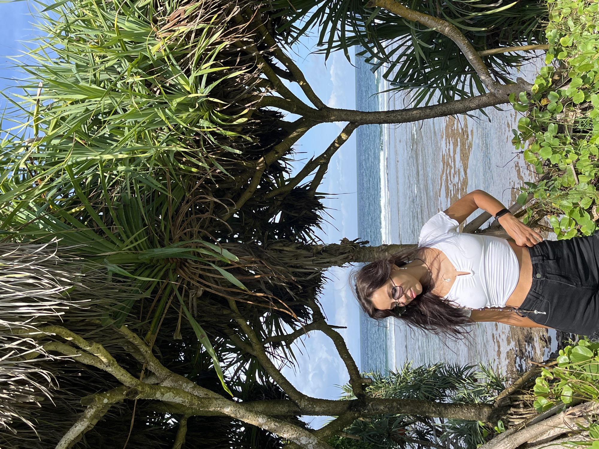 An outdoor, sunlit photograph captures a woman standing by a tropical coastline, framed by dense foliage. The image depicts a bright, clear day, likely midday or early afternoon, given the strong natural light.
The central subject is a woman with long, dark hair and sunglasses, wearing a white crop top and black jeans. She is adorned with a delicate necklace and a small decorative chain attached to her jeans. She stands with her body slightly turned, looking downwards with a relaxed expression, appearing to pose for the camera.
The setting is a vibrant coastal environment. Large, leafy green trees or shrubs, possibly pandan, dominate the foreground and sides of the frame, their branches and long, spiky leaves creating a natural arch or frame around the scene. Through the gaps in the foliage, the ocean is visible, featuring gentle waves breaking on a rocky and sandy shoreline. The sky above is a clear, light blue with scattered white clouds, enhancing the bright, tropical atmosphere. The overall scene evokes a sense of serene natural beauty in a warm climate, consistent with a location like Kuta Selatan, Indonesia.
No discernible text is visible in the image.