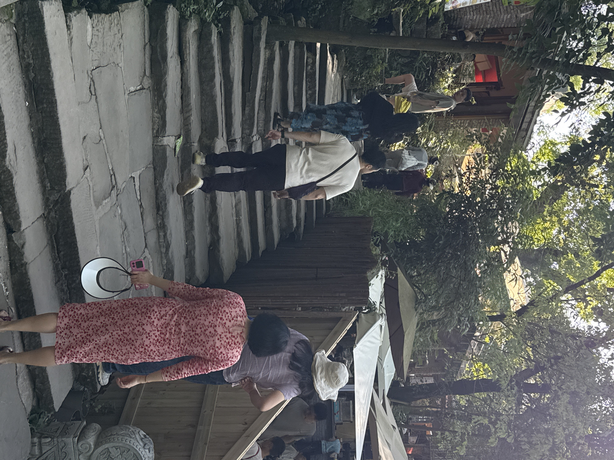The image shows an outdoor scene with multiple people walking on and around stone stairs. The setting appears to be urban, likely in Beibei, China, given the request.
People: Several individuals are walking up the stone steps, dressed in casual clothing. The variety of outfits suggests a tourist destination. One woman in a pink dress and holding a hat and phone.
Objects: Key objects in the image include the stone stairs, potentially leading to a temple or historical site. There are also trees, building structures, and some umbrellas nearby, hinting at shops or stalls in the area.
Actions: The primary activity is walking and sightseeing. The woman is taking a picture with her cell phone.
Environment: The presence of trees indicates a possible park or garden setting within the city. The light suggests it is daytime.