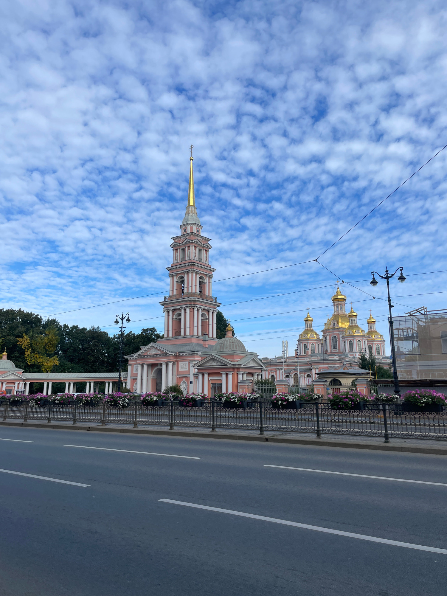 This is an eye-level, wide shot of the Smolny Cathedral in St. Petersburg, Russia, on a cloudy day. The pink and white Baroque cathedral dominates the center of the frame, with its tall, ornate bell tower and multiple golden domes. The cathedral is set behind a black metal fence adorned with planters filled with pink and white flowers. A wide asphalt road with white lane markings runs along the bottom of the image, indicating the presence of traffic. Trees line the background to the left of the cathedral, and modern buildings are visible to the right. Overhead, power lines crisscross the sky, and scattered, white, puffy clouds fill the blue sky. No people are visible in the image.