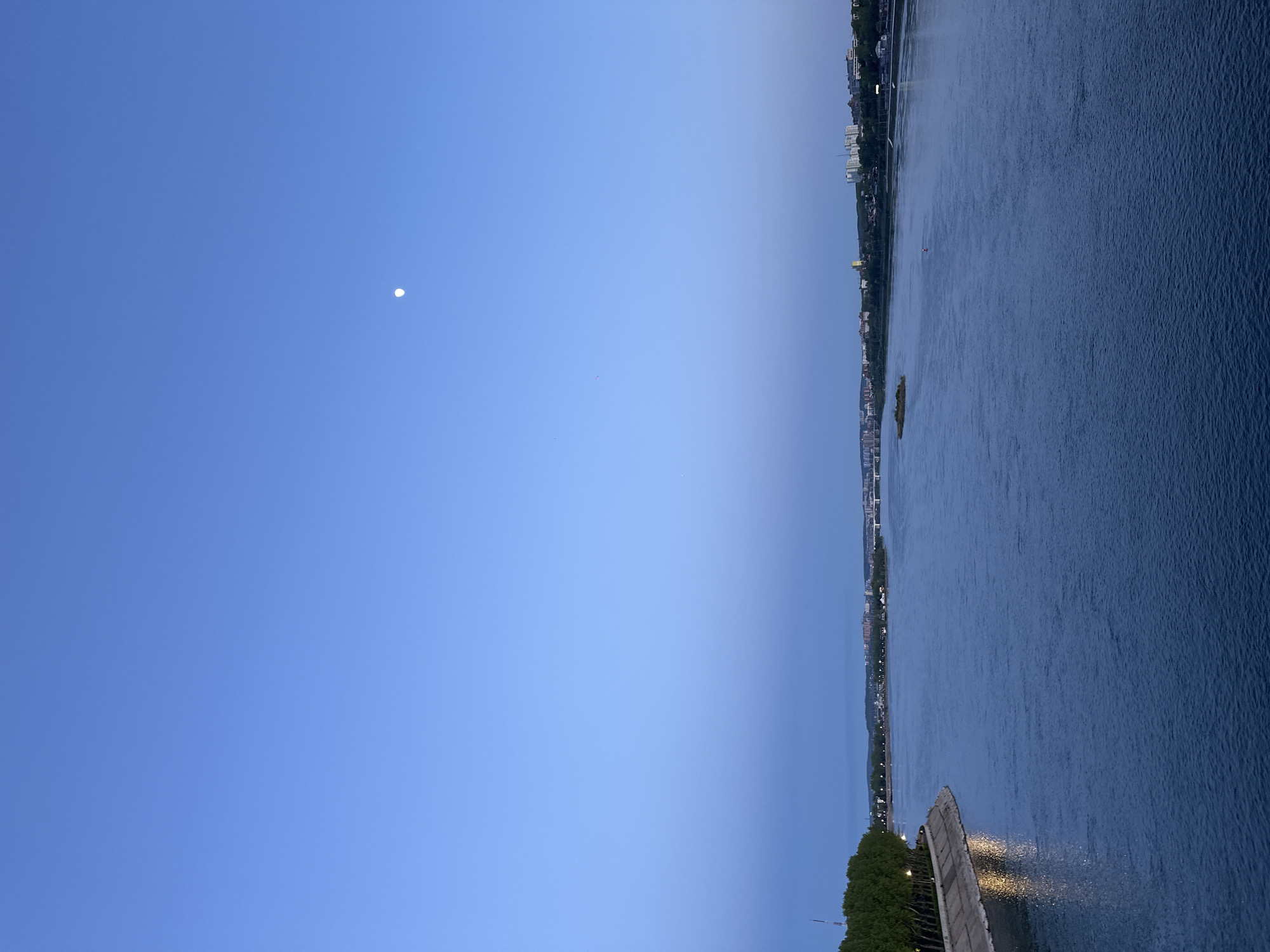 Captured at dusk, the image portrays the Angara River in Irkutsk, Russia. The river's dark blue waters fill the right half of the frame, with a city skyline visible along the opposite bank. A bright, almost full moon hangs in the upper left corner of the clear blue sky. A section of a bridge or embankment, lined with trees, appears in the lower right foreground. The lighting suggests early evening, with a gradual transition from the deep blue of the sky to the darker tones of the river.