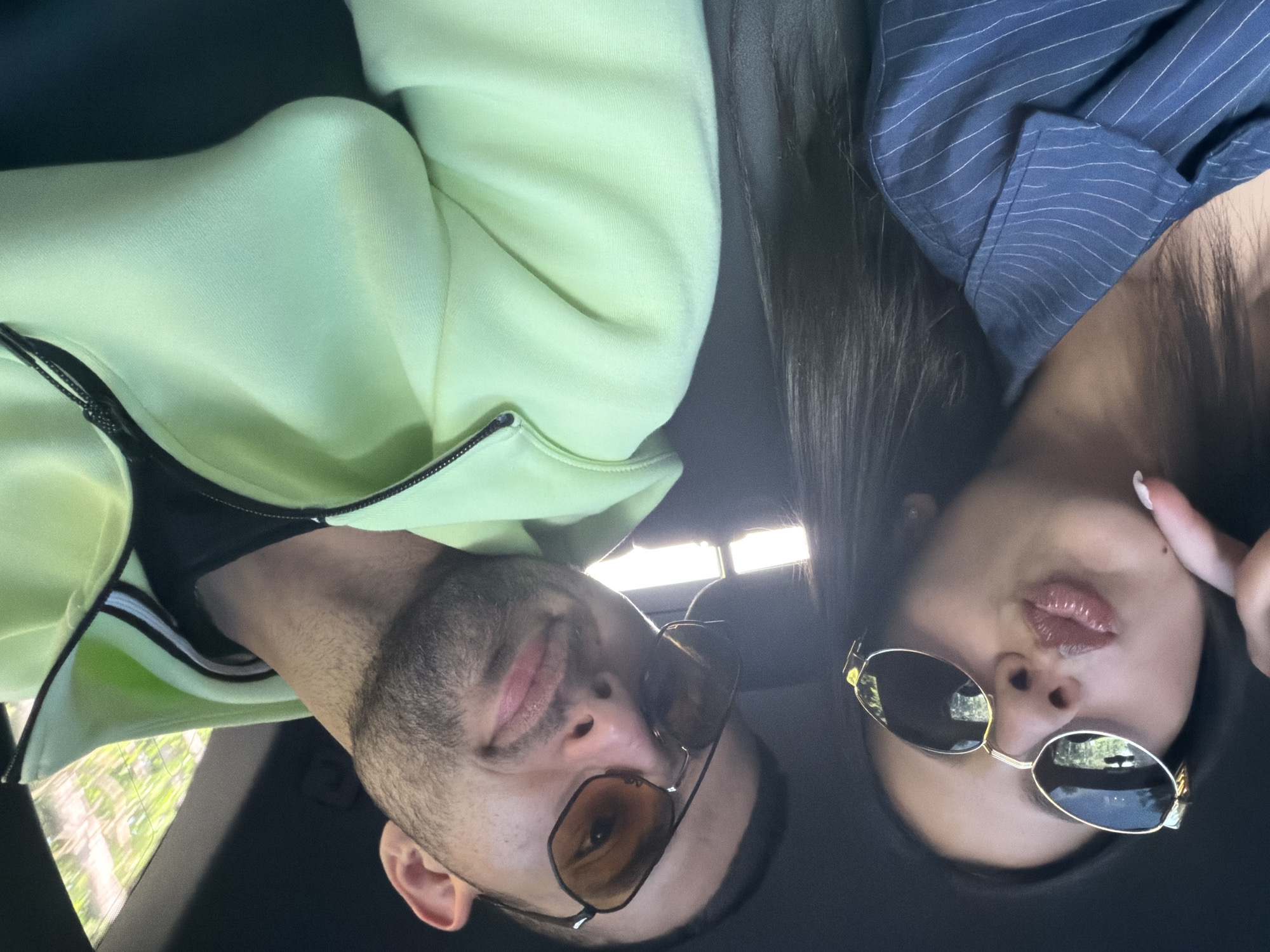 Two people are visible in a selfie taken inside a vehicle. On the left, a male figure wears a lime green zip-up sweatshirt, black t-shirt, and brown-tinted sunglasses. On the right, a female figure wears a pinstripe blue jacket, dark-tinted sunglasses, and has dark hair. The vehicle appears to be parked, with a view of foliage outside the window. The exact location in Obretenik, Bulgaria, cannot be determined from the image alone.