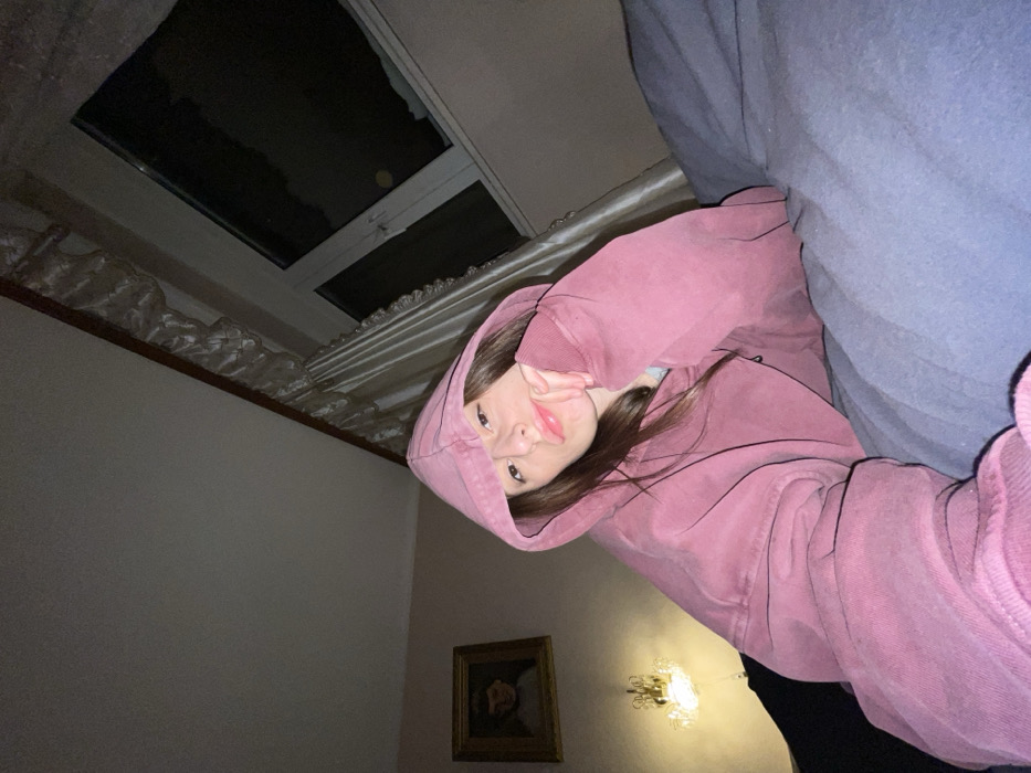 A young woman wearing a pink hooded sweatshirt looks at the camera. She has one hand near her face. The photo's perspective is angled upward, and a blue blanket is draped over the upper right corner. A rectangular window showing a dark sky is in the upper left, and the wall behind the woman is pale with a framed picture and illuminated by a decorative wall sconce.
