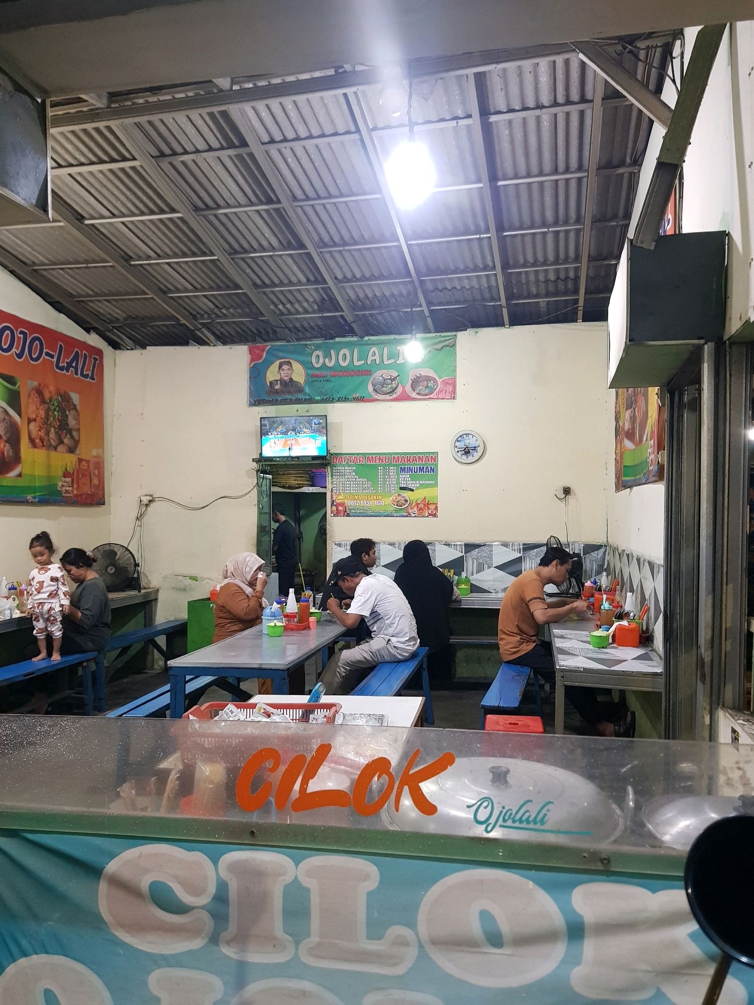 The image depicts the interior of a casual dining establishment, likely a street-side eatery or a small restaurant, in Setiamekar, Indonesia. The scene is illuminated by artificial lighting, casting a warm glow on the corrugated metal ceiling and the pale walls. Tables and benches, predominantly made of blue painted wood and metal, are arranged for seating.
Several individuals are present. In the foreground, a clear plastic barrier, possibly a display case or a protective shield, has large orange lettering that reads "CILOK" and smaller teal lettering that reads "Ojolali". Behind this, on the left, a young child stands on a blue bench, observing. Next to the child, an adult female sits at a table. Further into the scene, a woman in a brown headscarf is seated at a table, her hand near her face. To her right, a man wearing a light brown t-shirt is seated at another table, eating. Another person, partially visible, is seated at the same table. On the far right, a person wearing a white t-shirt is seated at a table. Another individual, a man with dark hair, is visible behind the counter area.
Various objects are scattered on the tables and surroundings, including plates, bowls, utensils, bottles of condiments, and a red plastic basket. A television screen mounted on the wall shows a sports broadcast, likely basketball, with Indonesian commentary. A clock with a blue face is also visible on the wall. Posters with text and images, including one with the text "OJOLALI" and a portrait, are displayed on the walls. The environment suggests a busy, functional space where food is prepared and served, with a focus on practicality and accessibility. The presence of a ventilation fan on the left side further indicates a functional food service setting. The overall impression is one of a lively, local dining spot.
