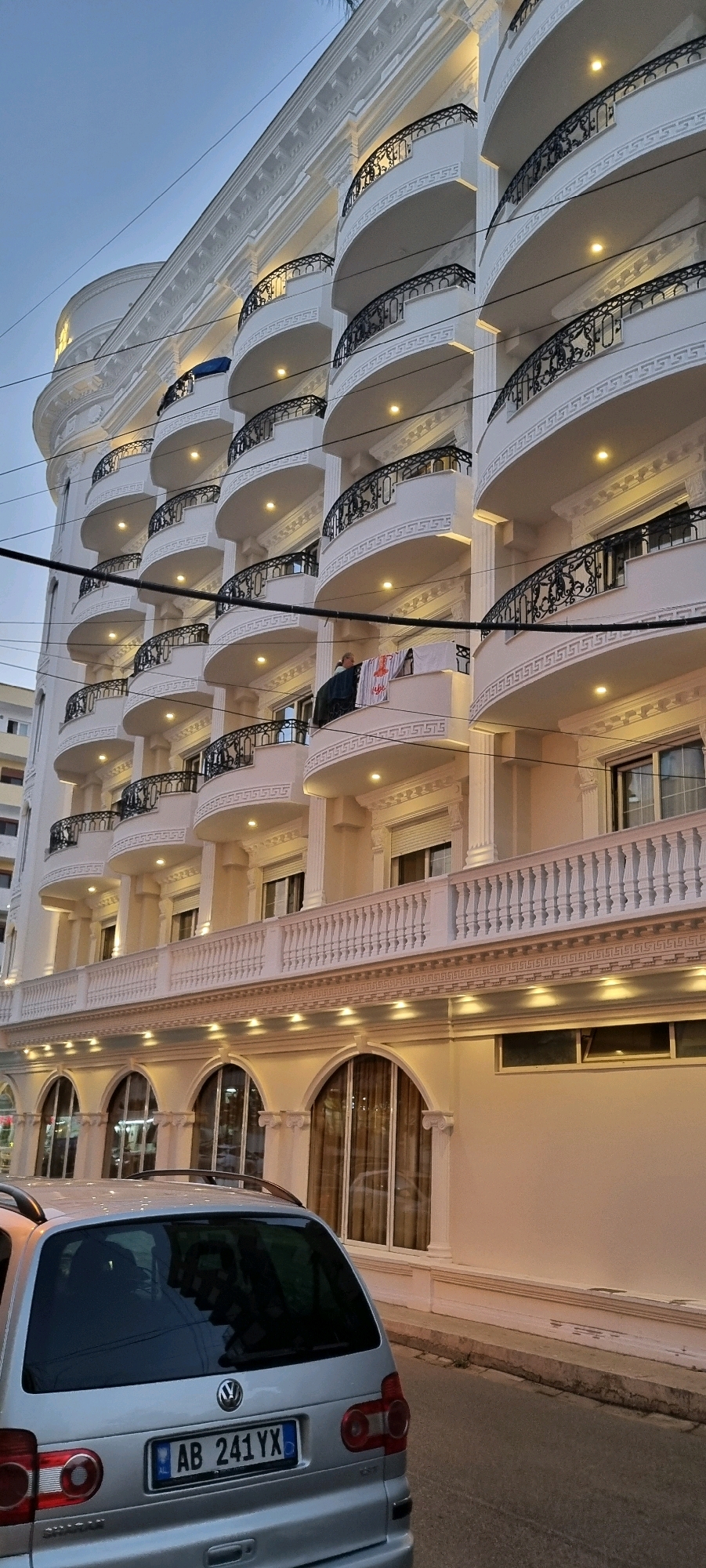 The image shows a multi-story white building with a modern architectural style, characterized by curved balconies with black wrought-iron railings and ornate white trim. The building is illuminated by warm-toned lights, suggesting it is either dusk or evening. Several windows with rolling shutters are visible on different floors. A person is standing on one of the balconies, with laundry hanging on a railing. In the foreground, the rear of a silver Volkswagen Sharan station wagon is visible, parked on a street in front of the building. The license plate of the car reads "AB 241 YX". The street surface appears to be asphalt, with a concrete curb along the edge of the sidewalk. The sky above is a clear, pale blue, indicating it is likely early morning or late evening. The building's design suggests it could be a hotel or residential complex. The location is identified as Bashkia Durrës, Albania.