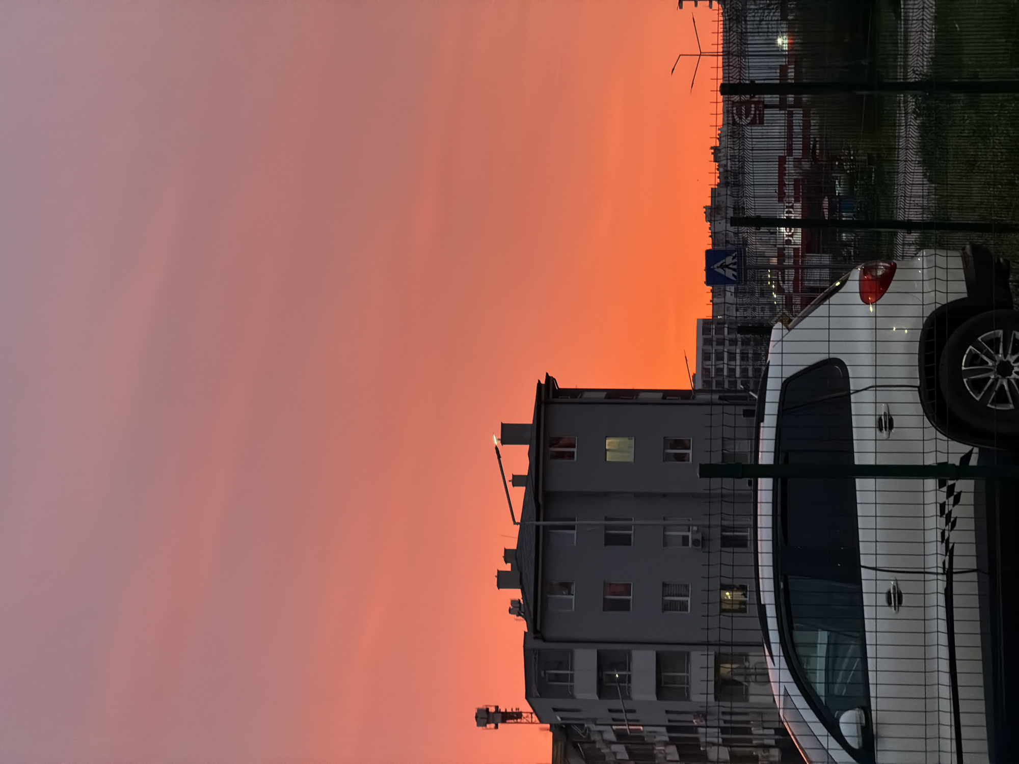 The image shows a sunset in an urban area, possibly Varna, Bulgaria, given the user's prompt. The sky exhibits a gradient from pink to orange. There is a multi-story building in the mid-ground. A white car is parked near a chain-link fence. A pedestrian crossing sign is visible. Other buildings are in the background. A street light is on near the top of the building.