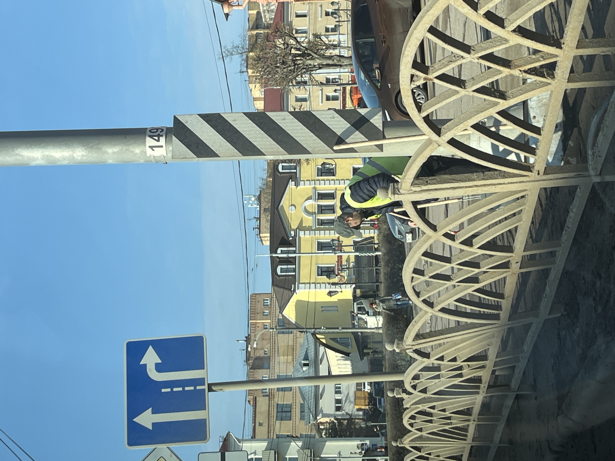 The image, likely taken from inside a vehicle, shows a street scene in what is reported as the city of Klin, Russia. A traffic sign on the left indicates a lane division ahead with a straight or right turn available from the right lane and a left turn from the left lane. Next to the traffic sign is a support post that has black and white diagonal stripes near its top, with a numbered label "149" attached to it. In the background are various multi-story buildings. A worker wearing a safety vest is seen working on an elaborate metal fence that is close to the camera, obscuring some of the background. A brown car is also visible parked behind the fence. The sky is blue.