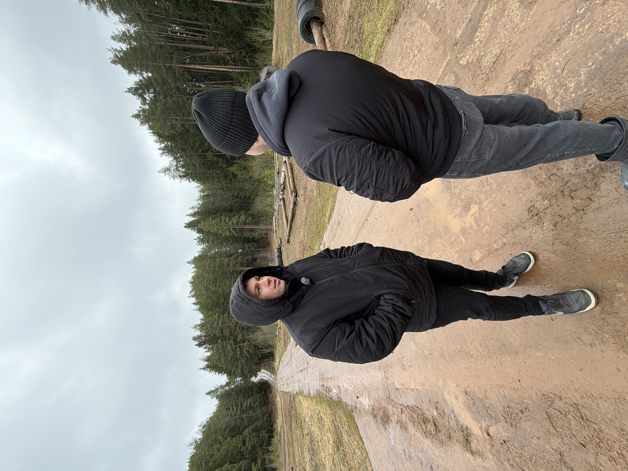 Two men stand on a muddy path in what appears to be a wooded area. One man is facing the camera, wearing a black hooded jacket and black pants with grey shoes. The other man, further up the path, wears a black beanie, a dark jacket, and grey jeans. Trees line the background under an overcast sky. According to metadata, the image was taken in Прикамский, Russia.