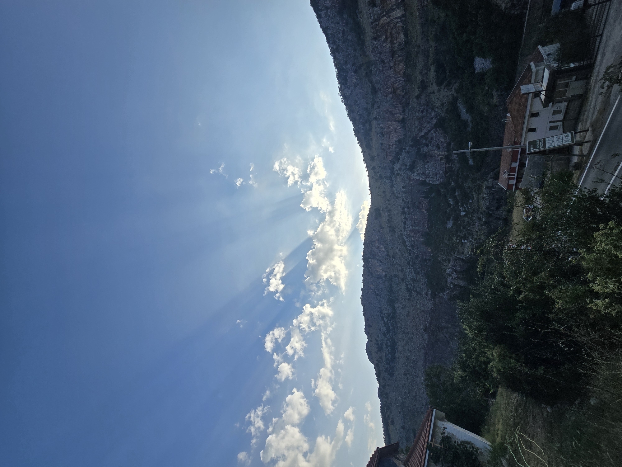 A wide-angle, vertical shot depicts a mountainous landscape under a clear blue sky. The sun, positioned behind a ridge of clouds, casts prominent crepuscular rays across the sky, illuminating patches of clouds. The rugged, rocky mountain slopes dominate the upper right and central background, sparsely dotted with trees. In the foreground and mid-ground, a few houses with red-tiled roofs are visible. One house on the left has white walls and is partially covered in ivy. Another building complex with white walls and red trim is situated closer to a road in the right mid-ground. A signpost with various pictograms and text, including "AUTO CAMP" and "TITOGRAD", stands near this complex. Lush green vegetation, including trees and grass, fills the areas around the houses and on the lower slopes. The road curves through the scene in the lower right. The overall scene suggests a rural or semi-rural setting in a mountainous region, potentially near Podgorica, Montenegro, given the "TITOGRAD" inscription on the sign, which was the former name of Podgorica.