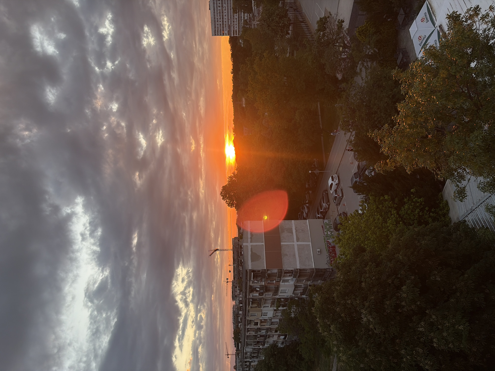 The image shows a sunset over the city of Plovdiv, Bulgaria. A large apartment building with construction cranes is visible, as are trees, a parking lot, and other buildings. The sky is filled with clouds that are illuminated by the setting sun. The scene is likely taken from an elevated vantage point.