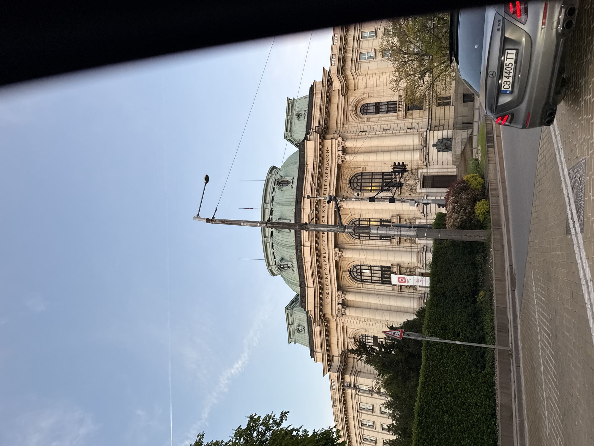 The image shows the front of the Saint Kliment Ohridski University of Sofia in Sofia, Bulgaria. The building is large and ornate, with a pale facade and green copper roofs. The name of the university is written in Cyrillic along the front. A streetlight stands in front of the building. A gray car is parked along the street, with its license plate visible. A portion of the car is outside the frame. Green hedges line the sidewalk. A triangular road sign is present near the hedge. The sky is blue and clear. The image appears to be taken from inside a vehicle, as a black area obscures the upper left.