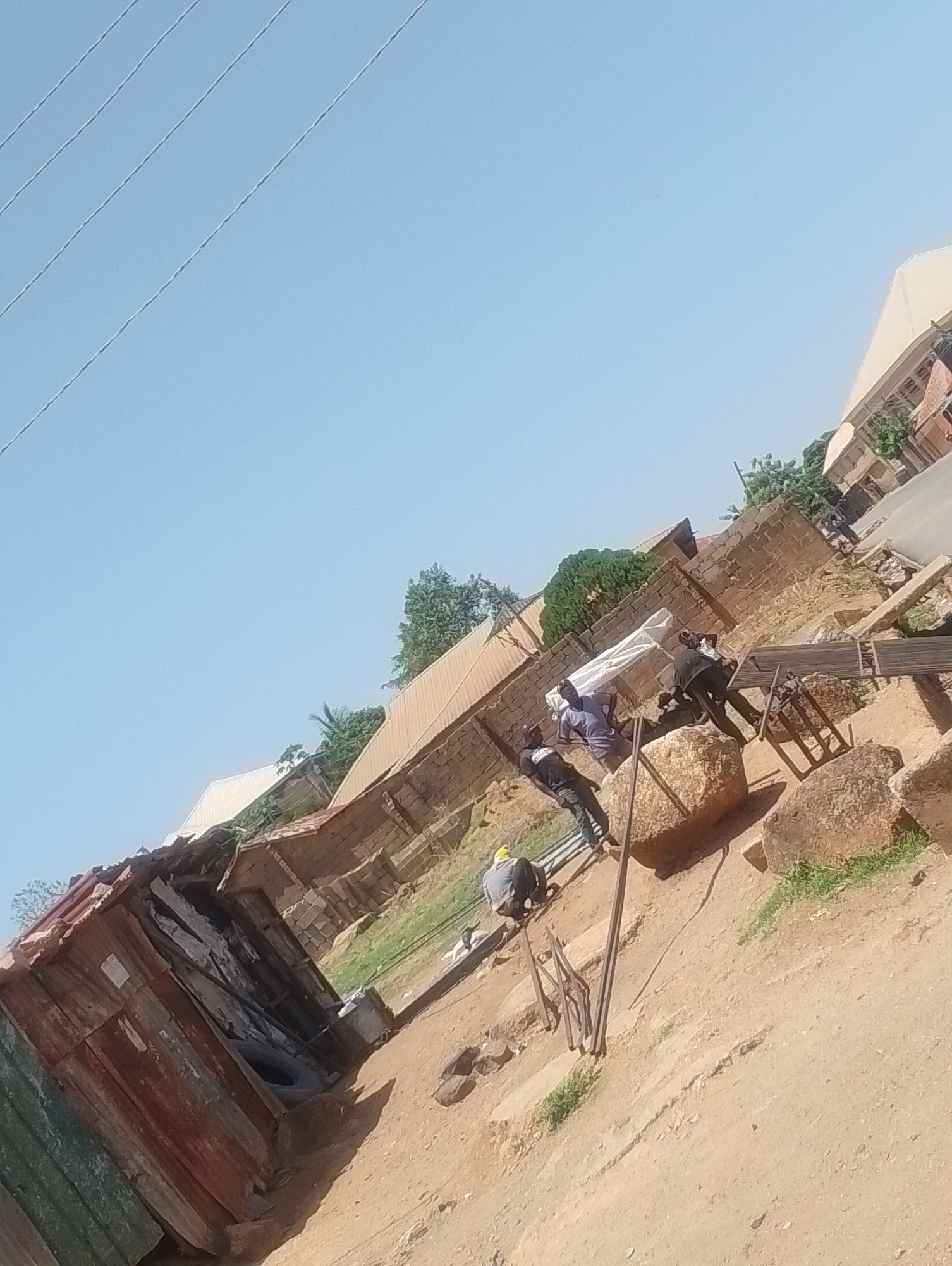The image depicts a roadside scene in Jos, Nigeria. Three men are working on construction-related tasks amidst exposed dirt. One man wears a yellow hard hat and is crouching, while two others are interacting near a large rock. Metal bars are scattered near them. A rundown wooden structure with corrugated iron sheets stands to the left of the frame. Behind the men, a brick building with a metal roof is visible. Further back, a road with more buildings along the roadside is seen. Overhead power lines are present in the upper left corner of the image. The sky is clear.