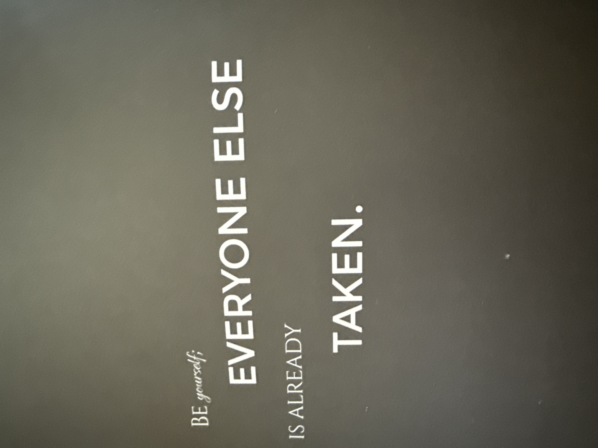 The image shows a poster with the text "BE yourself; EVERYONE ELSE IS ALREADY TAKEN." The background of the poster is a dark gray or black. The text is in white. The image is taken indoors with artificial light. The location of the poster in the city of Troyan, Bulgaria, cannot be determined from the image.