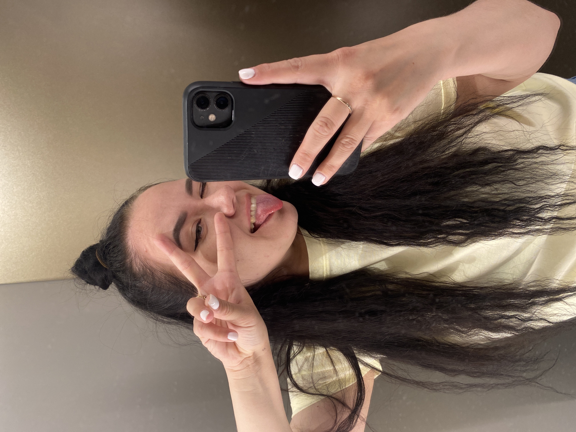 A woman is taking a selfie with her smartphone. She is wearing a light yellow top and has long dark hair. She is making a peace sign with one hand and holding the phone with the other. Her nails are painted white. She has one eye closed and her tongue is sticking out. The background appears to be a wall with a change in color or material. The location is the city of Perm, Russia.