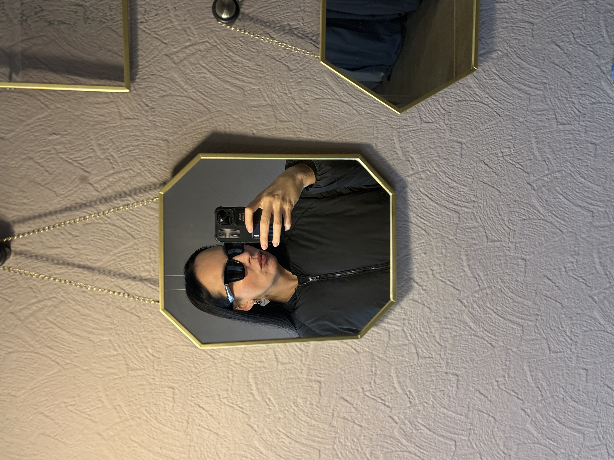 A woman takes a selfie in a wall-mounted, octagon-shaped mirror with a gold frame. The mirror is attached to the wall by a gold chain connected to a knob. The woman has dark hair and wears sunglasses and a black jacket. Her phone case shows a pair of eyes. Other mirrors with gold frames are also visible. The wall is beige with a textured pattern. It is not possible to identify the location as Chaikovsky, Russia based on the image.