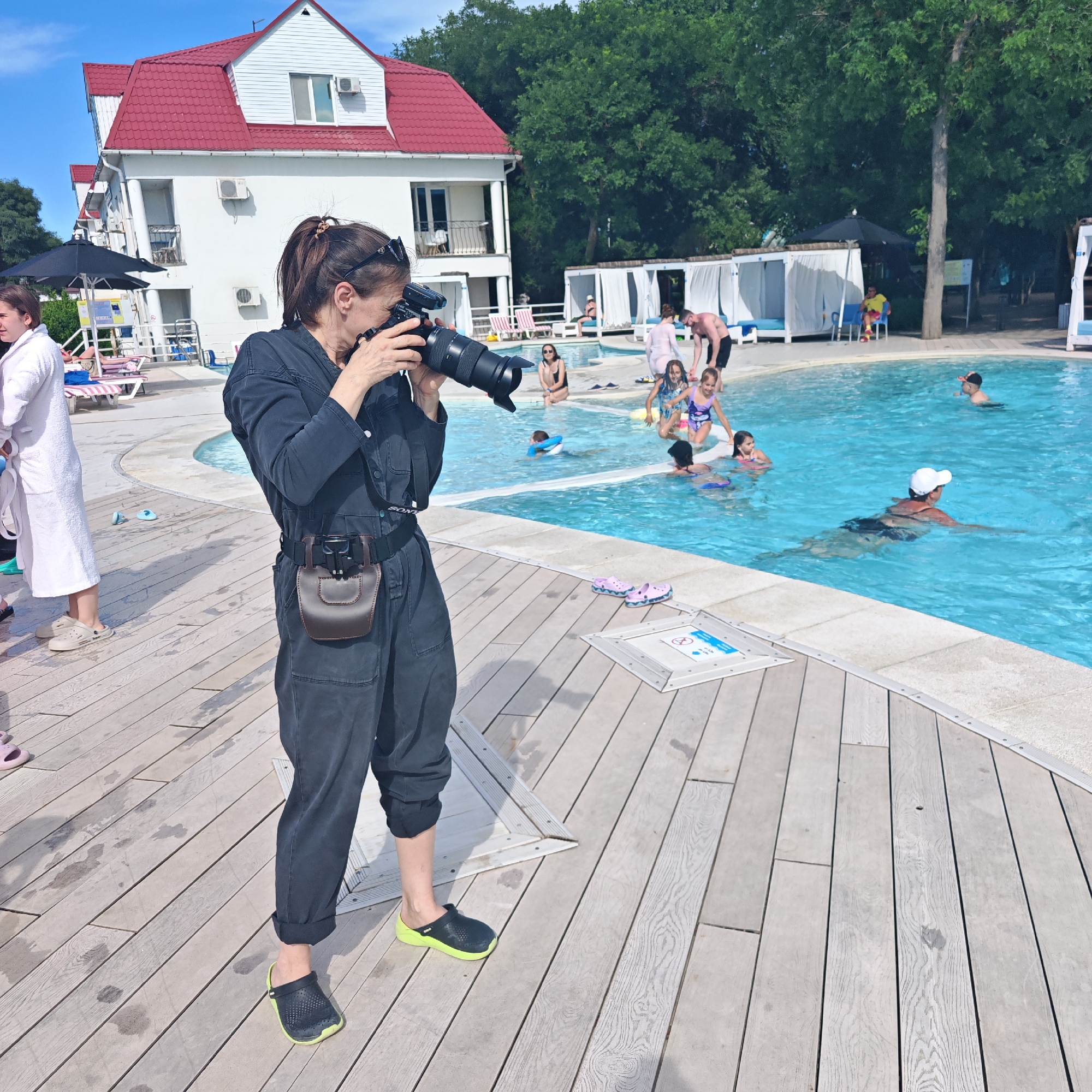 In the city of Shchyolkino, Ukraine, a woman is positioned on a wooden deck next to a swimming pool, actively photographing a group of people. She is wearing a dark jumpsuit and has a camera with a long lens in her hands, focused on the pool area. A brown leather pouch is attached to her belt. Around the pool, several individuals are visible. In the foreground to the left, another person in a white robe is standing. The background features a white building with a red roof and lush green trees. The swimming pool is filled with blue water, and several children and adults are enjoying themselves. Some are swimming, while others are sitting or standing near the water's edge. White cabanas are set up around the pool area, suggesting a resort or leisure facility. The wooden deck has a slightly wet appearance, with some footprints visible, indicating recent activity. The overall scene depicts a lively summer day at a pool.