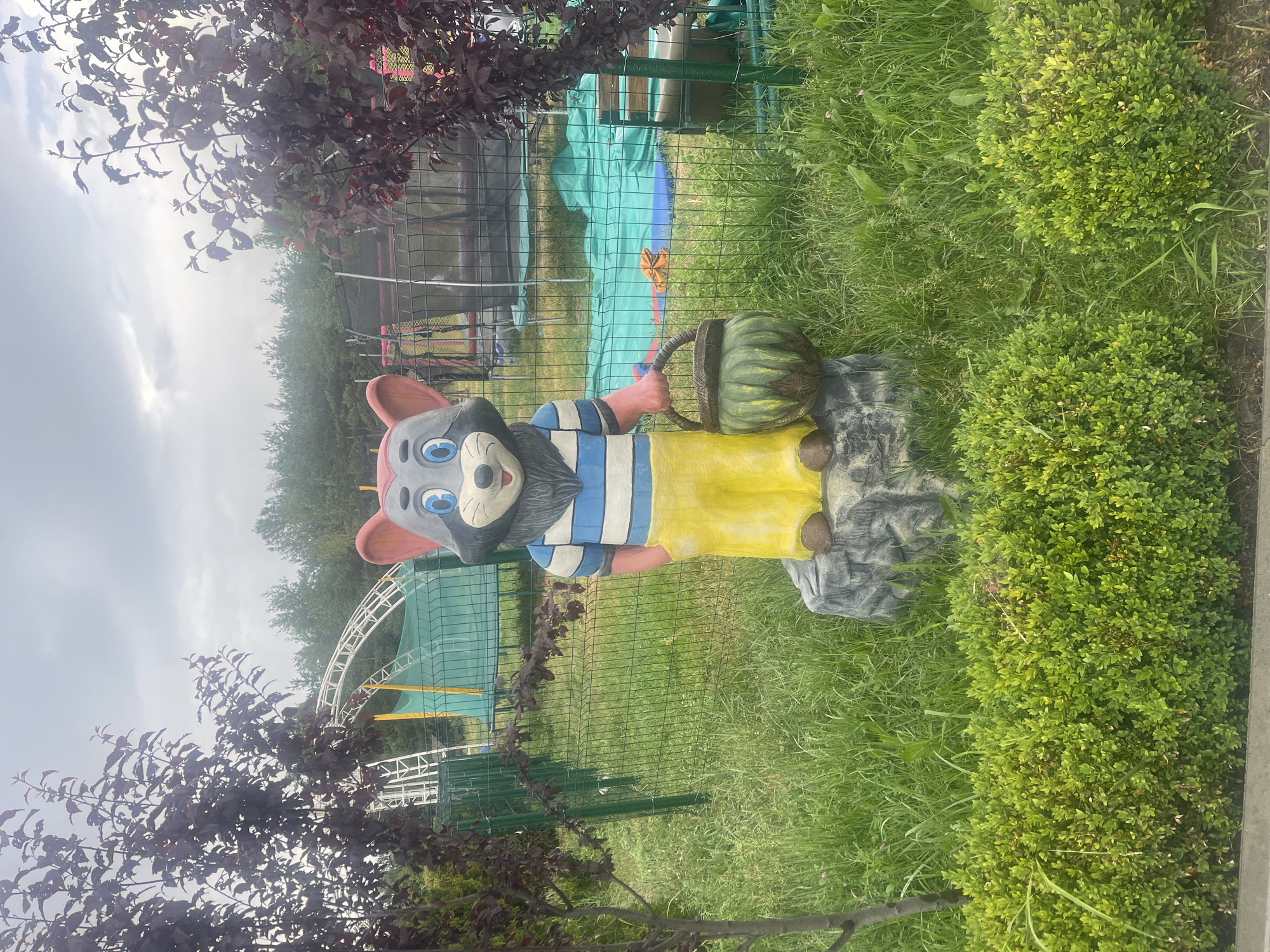 An outdoor, medium-shot image features a cartoonish mouse statue standing on a gray rock base amidst lush green grass and foliage. The statue is depicted with a gray body, red ears, blue eyes, and a friendly expression. It wears a blue and white striped shirt and yellow pants, and holds a basket containing what appears to be a pumpkin or gourd in its left hand. Behind the statue, a green wire fence encloses an area with a white roller coaster structure, a trampoline, and green tarps covering some equipment. Dark-leaved trees flank the sides of the frame, and a backdrop of trees and an overcast sky completes the scene. The setting appears to be an amusement park or recreational area.