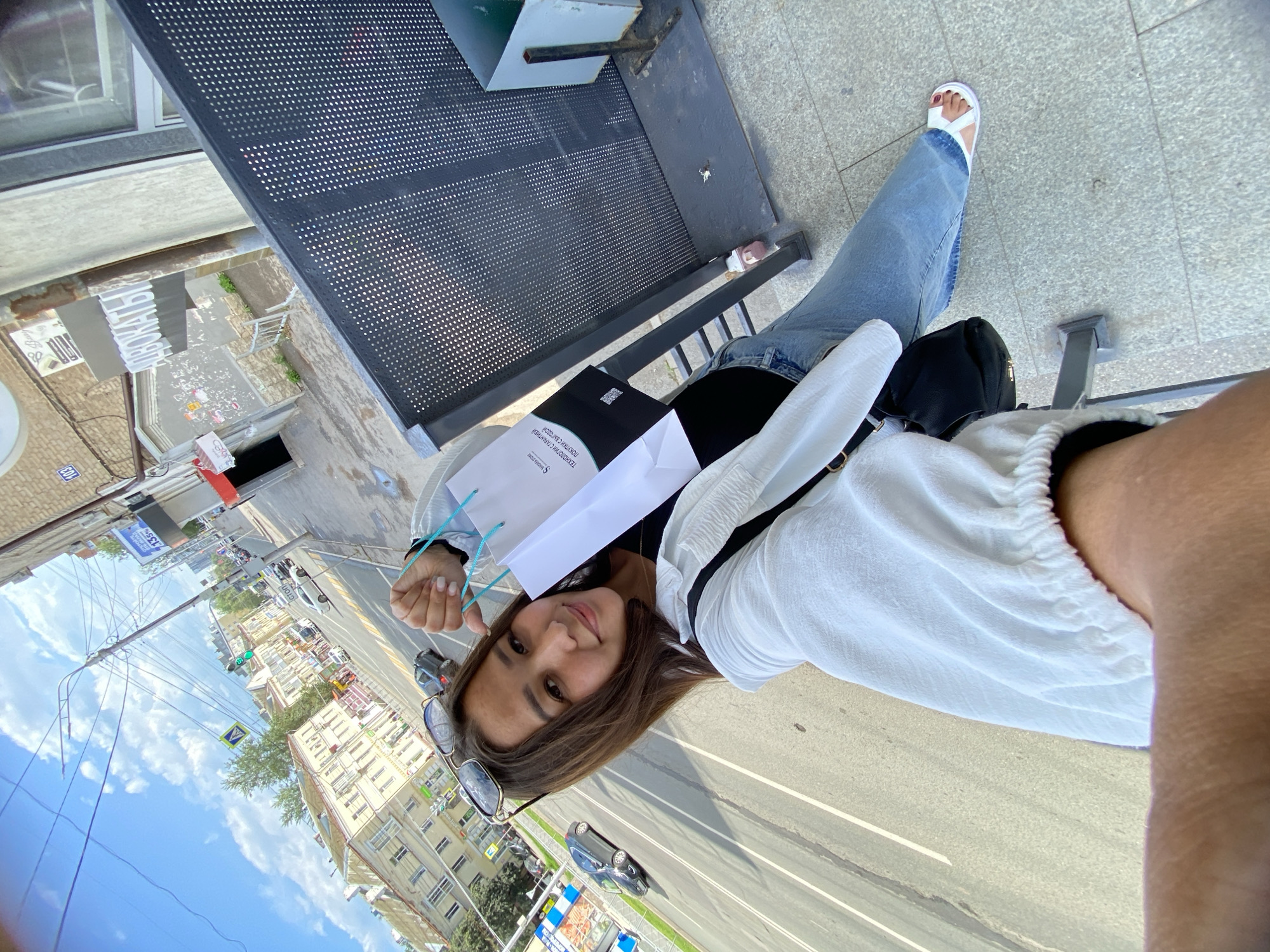A young woman with brown hair, wearing a white open-neck shirt, blue jeans, and white sandals, stands on a sidewalk in front of a building. She is holding a white shopping bag with blue handles. She is looking at the camera and smiling. The background shows a street with cars, buildings, and a blue sky with white clouds. A sign on the building behind her has Russian text. The overall scene suggests a casual day out in a city. The specific location is in Ufa, Russia, with the street name "Lenina" visible on a street sign.