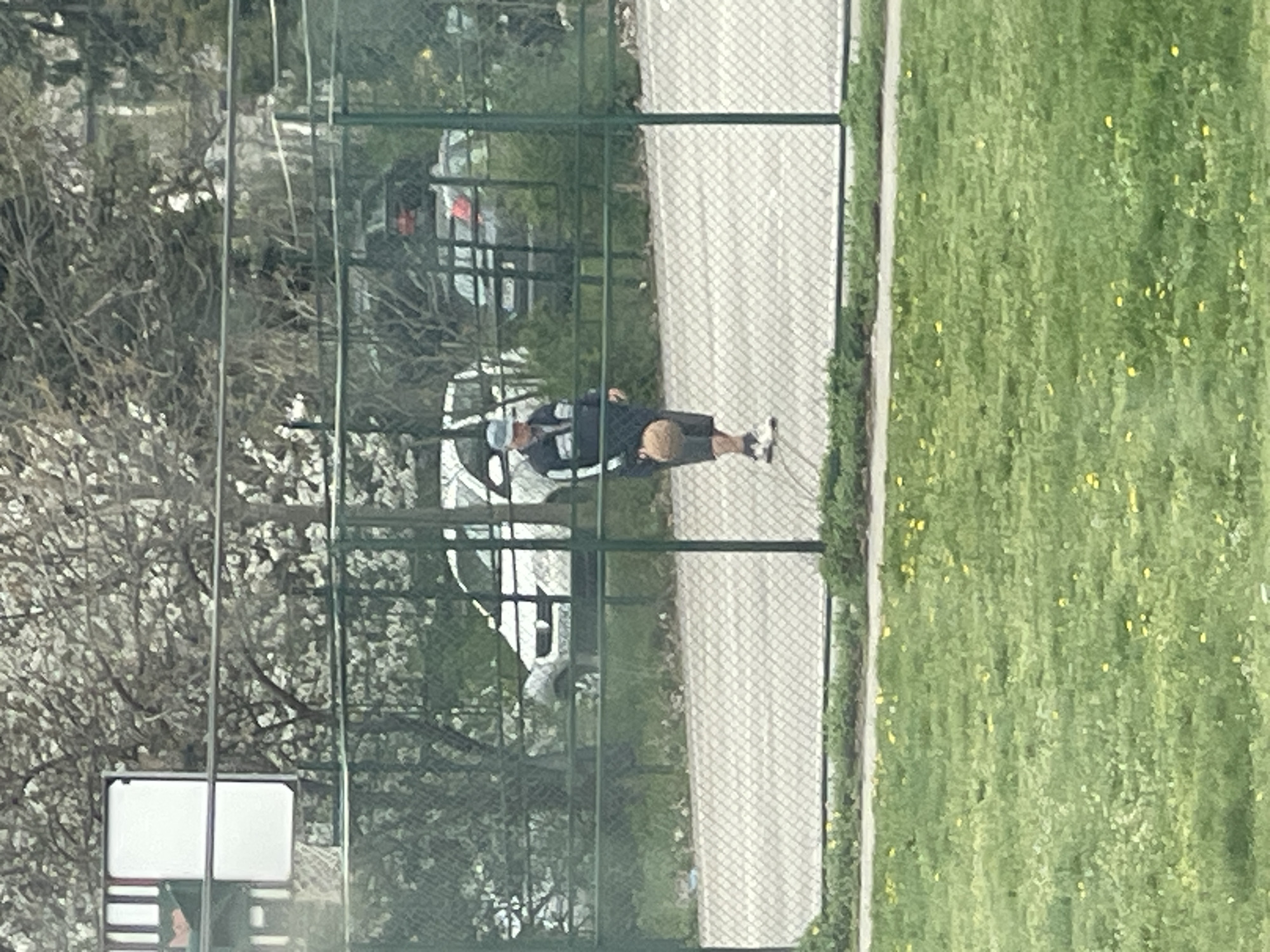 A medium shot depicts a man walking on a gray path beside a green grassy area. A fence separates the path and grassy area from the foreground. The man is seen wearing a black top, shorts, a baseball cap, and walking shoes. Behind the fence, there are bare trees with white blooms, parked cars, and possibly residential buildings. The photograph was taken through a fence with a green frame. The location is possibly in a park in Sofia, Bulgaria. There is a backboard behind the fence.