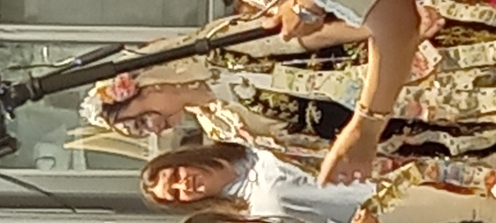 The image shows a group of people outdoors, with a focus on a woman in traditional attire. She is wearing a patterned dress adorned with what appear to be paper money. She has flowers in her hair and is wearing jewelry. A woman to her left is wearing a light blue shirt. The background suggests an outdoor setting, possibly in a town square or street, with architectural elements visible. There is a tripod and camera in the background, indicating that the scene is being filmed or photographed. The overall impression is of a celebratory event, possibly a wedding or festival. There is no specific information about the location in Lyuben, Bulgaria.