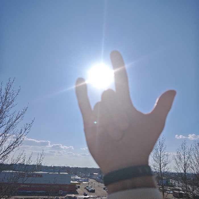 The image presents an elevated, wide-angle view over a city landscape in Rostov Veliky, Russia, with the sun positioned centrally in the upper frame. In the immediate foreground, a human hand, out of focus, is raised, forming a "rock on" gesture with the index and pinky fingers extended. Two bracelets, one dark and one lighter, are visible on the wrist, along with the cuff of a light-colored garment. The sun, bright and causing significant lens flare, is set against a clear blue sky with a few scattered white clouds near the horizon. In the mid-ground, a large, multi-story building featuring a red base and a prominent white upper facade with the Cyrillic text "ЭКСТРА" (EXTRA) is clearly visible. This building is identified as the EXTRA hypermarket at Okruzhnaya Ulitsa, 19. A parking lot with numerous vehicles occupies the space in front of this building, and patches of snow are visible on the ground. Bare, deciduous tree branches frame the left and right edges of the image, indicating a late winter or early spring season. The background shows a continuation of the urban environment with other distant buildings under a bright sky.