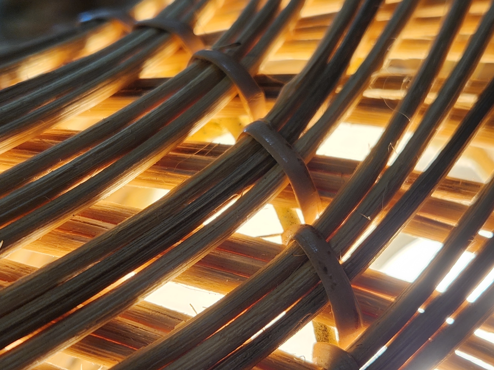 This is a close-up, angled shot of woven rattan. The rattan is arranged in a criss-cross pattern, with thicker, darker strands alternating with thinner, lighter strands. The lighter strands are illuminated from behind, creating a warm, glowing effect. The rattan is tied together in places with a darker, thinner material. The overall impression is one of texture and light. The location is an unknown interior in Maitland, United States of America.