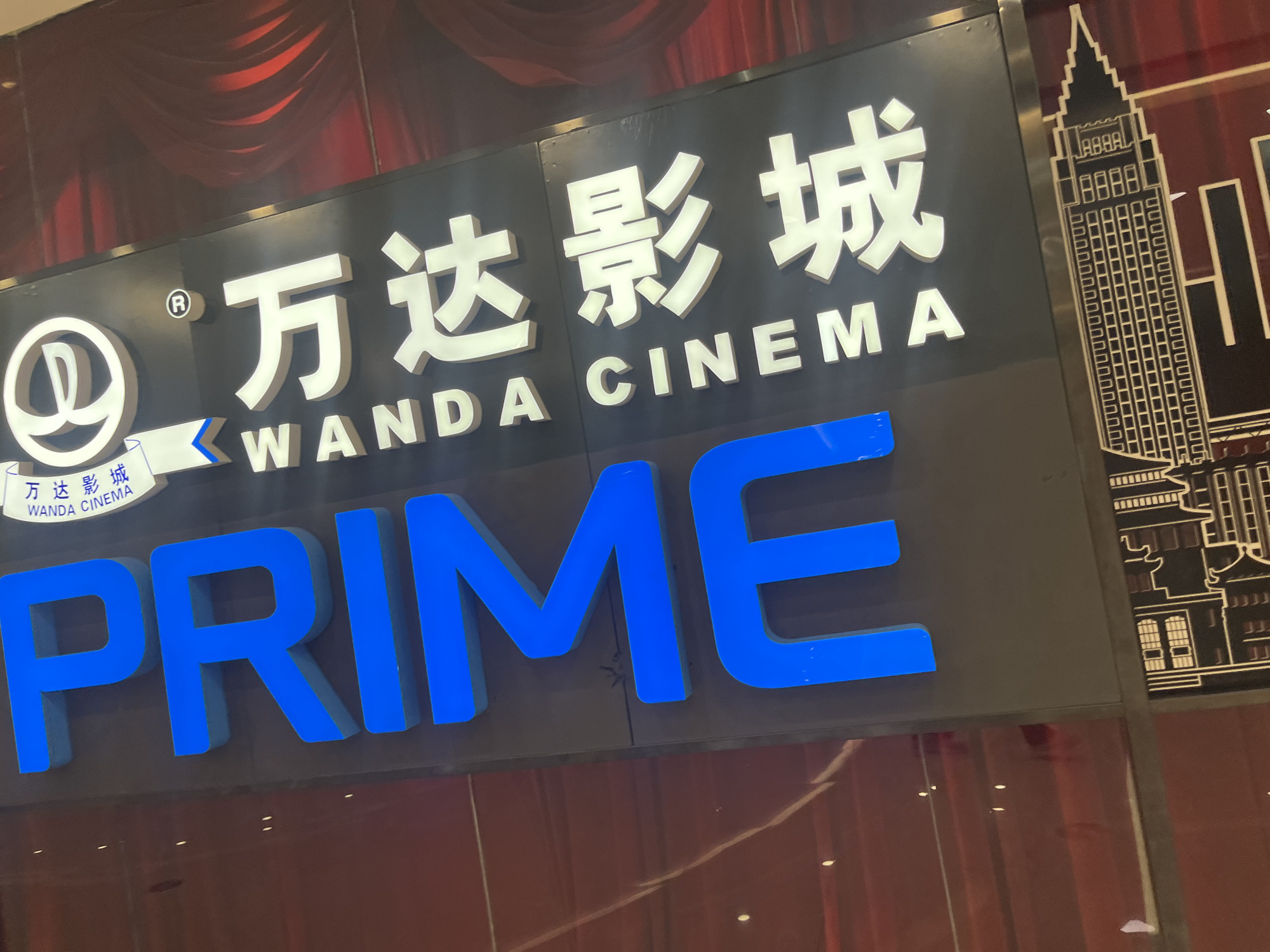 The image shows a close-up of a bright blue "PRIME" sign in large, blocky letters. Above it, a black sign displays white Chinese characters and the text "WANDA CINEMA" with a logo featuring a stylized "W" inside a circle. The background features a dark, textured surface with red drapery-like elements and an illustration of a stylized skyscraper. This signage is likely located in the Shapingba District of China.