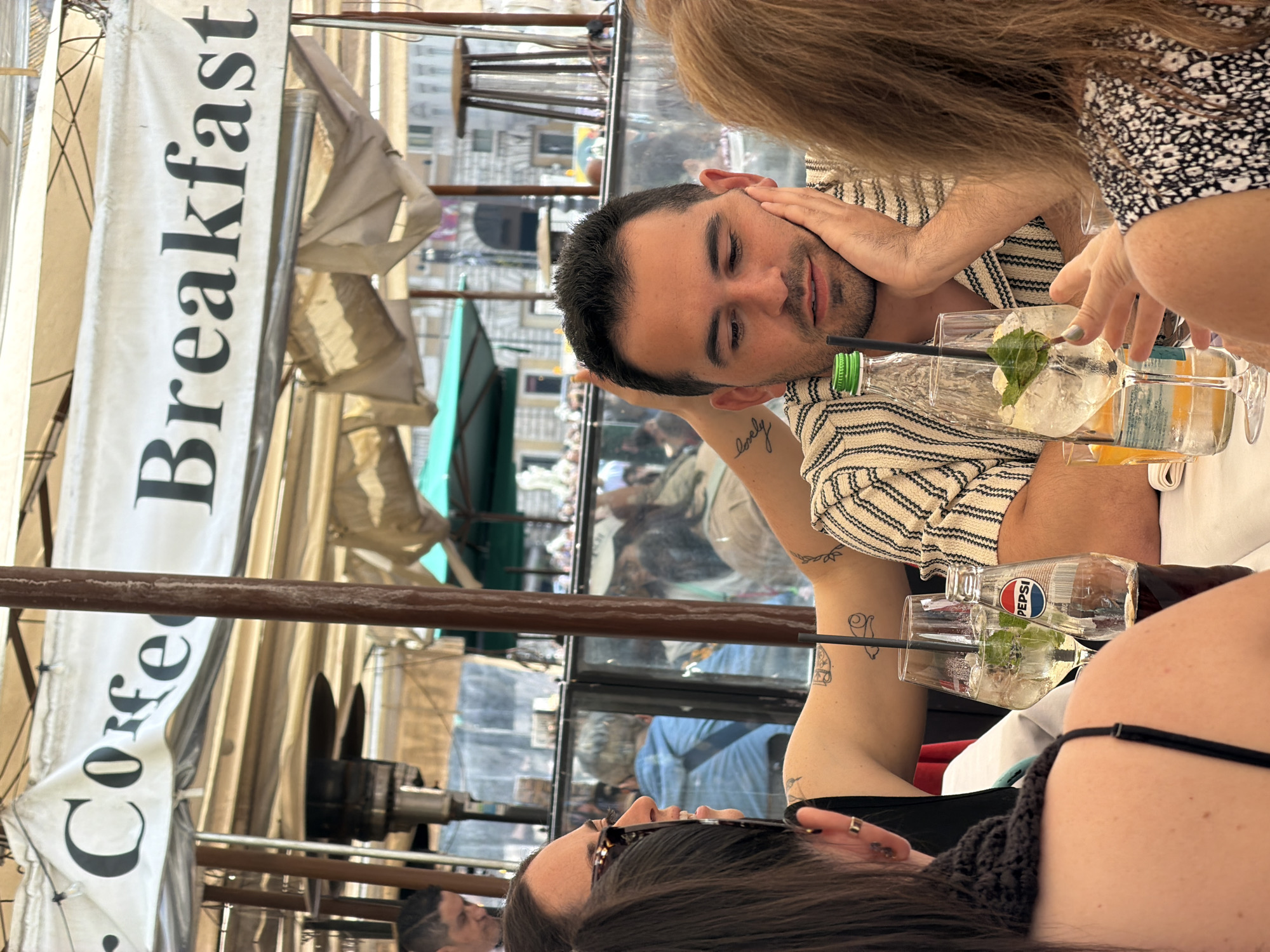 The image captures an outdoor cafe scene, likely in Rome, Italy, during the daytime, with several people seated around a table. The perspective is tilted, showing the subjects and surroundings as if viewed from an elevated, slightly rotated angle.
In the central foreground, a man with short dark hair and a beard is visible, wearing a striped shirt. He appears relaxed, resting his head on his hands. A tattoo reading "lovely" is distinctly visible on his forearm, along with other small, abstract tattoos further up his arm. To his right (viewer's left in an upright orientation), the upper body and long blonde hair of another woman are partially visible, her hand resting near the man's head. On the opposite side of the table, facing away, is a woman with long dark hair, wearing a black strap top, with small tattoos visible on her arm.
On the table are several drinks, including glasses filled with ice, mint leaves, and clear liquid, possibly cocktails or infused water. A clear plastic bottle with a green cap and a black straw, along with a Pepsi bottle, are also present. Other glasses contain an amber-colored beverage.
The setting appears to be an outdoor seating area of a cafe or restaurant, indicated by the awnings and structures above. A prominent white banner hangs vertically on the left side of the frame, displaying the text "Coffee Breakfast" in black. In the background, visible through a glass partition, are reflections of what appears to be classic European architecture and other people, suggesting a bustling urban environment. The bright lighting indicates it is daytime.