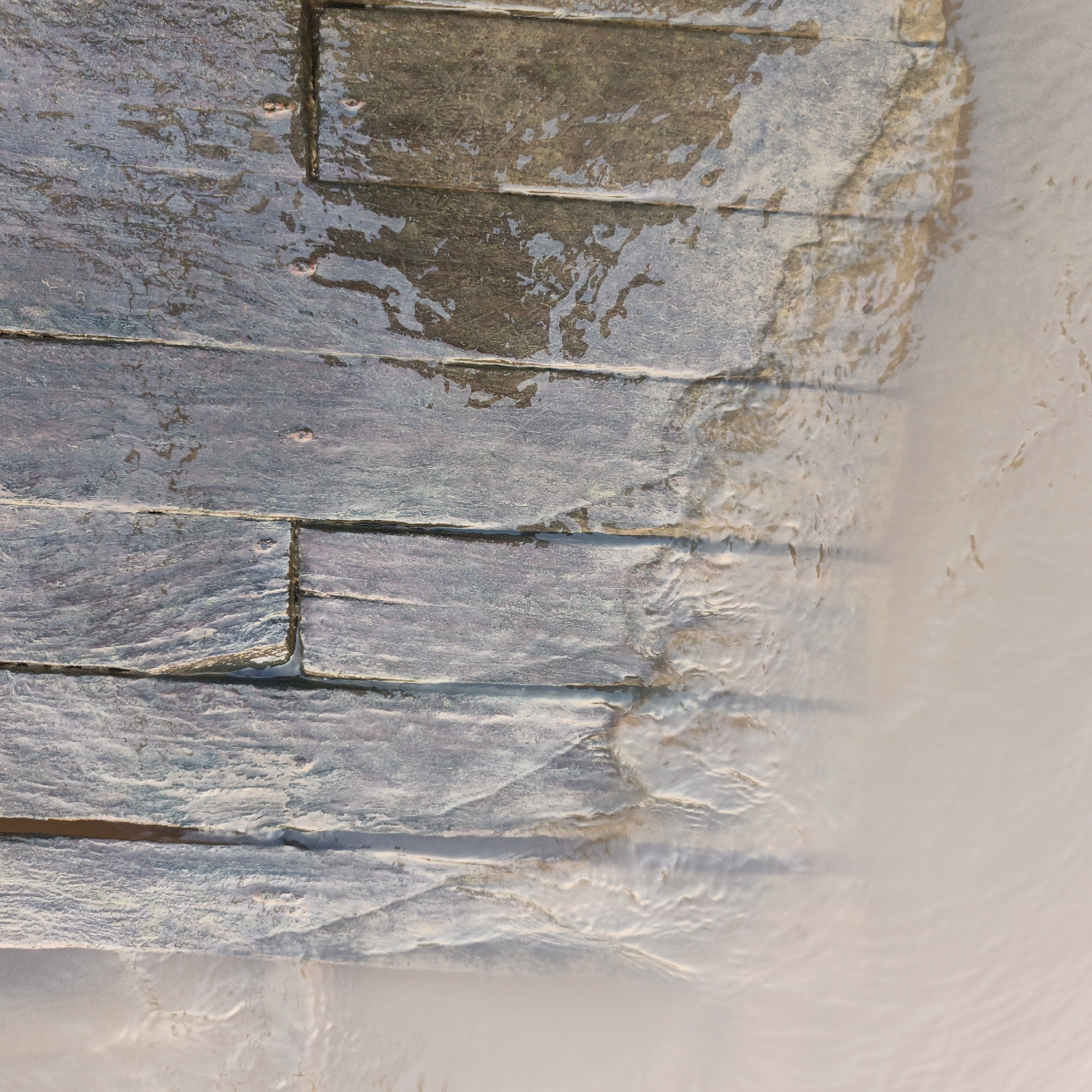 This is a close-up, top-down, slightly angled, forensic photograph of a damp, textured stone wall that appears to be in Sungai Rasau, Indonesia. The wall is constructed of rectangular, horizontally oriented stone blocks, with visible grout lines between them. The stones exhibit variations in color, ranging from dark grey and brown to lighter bluish-grey tones, with a rough, weathered surface. Water is present on the surface of the wall, creating reflective patches and streaks, particularly on the upper and mid-sections. These water patterns suggest recent exposure to moisture, possibly from rain or mist. The right side of the image transitions into a softly focused, lighter colored area, which could be a lighter colored surface, fog, or water. There are no people or animals visible in the frame. The overall impression is one of a textured, wet outdoor wall surface.