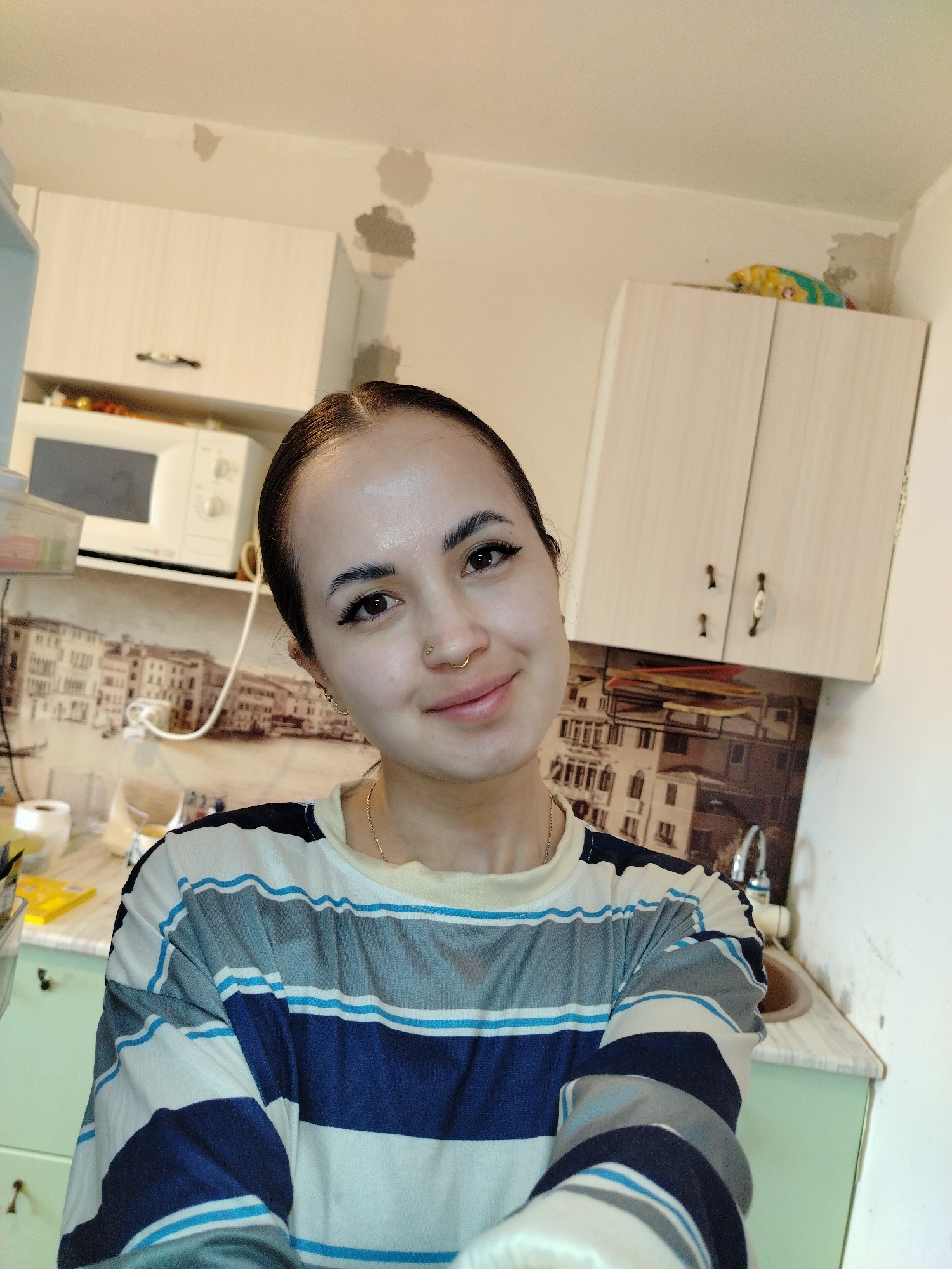 A medium-shot portrait depicts a young woman in a kitchen. She has dark hair pulled back from her face, dark eyebrows, eyeliner, and a nose ring. She is wearing a striped, long-sleeved shirt with shades of blue, gray, and white. The background shows kitchen cabinets, a microwave, and a print depicting a city with buildings, suggestive of an Italian setting. The cabinets are light-colored wood, and some areas of the wall appear to have patches or repairs. The location is Noviy, Russia.