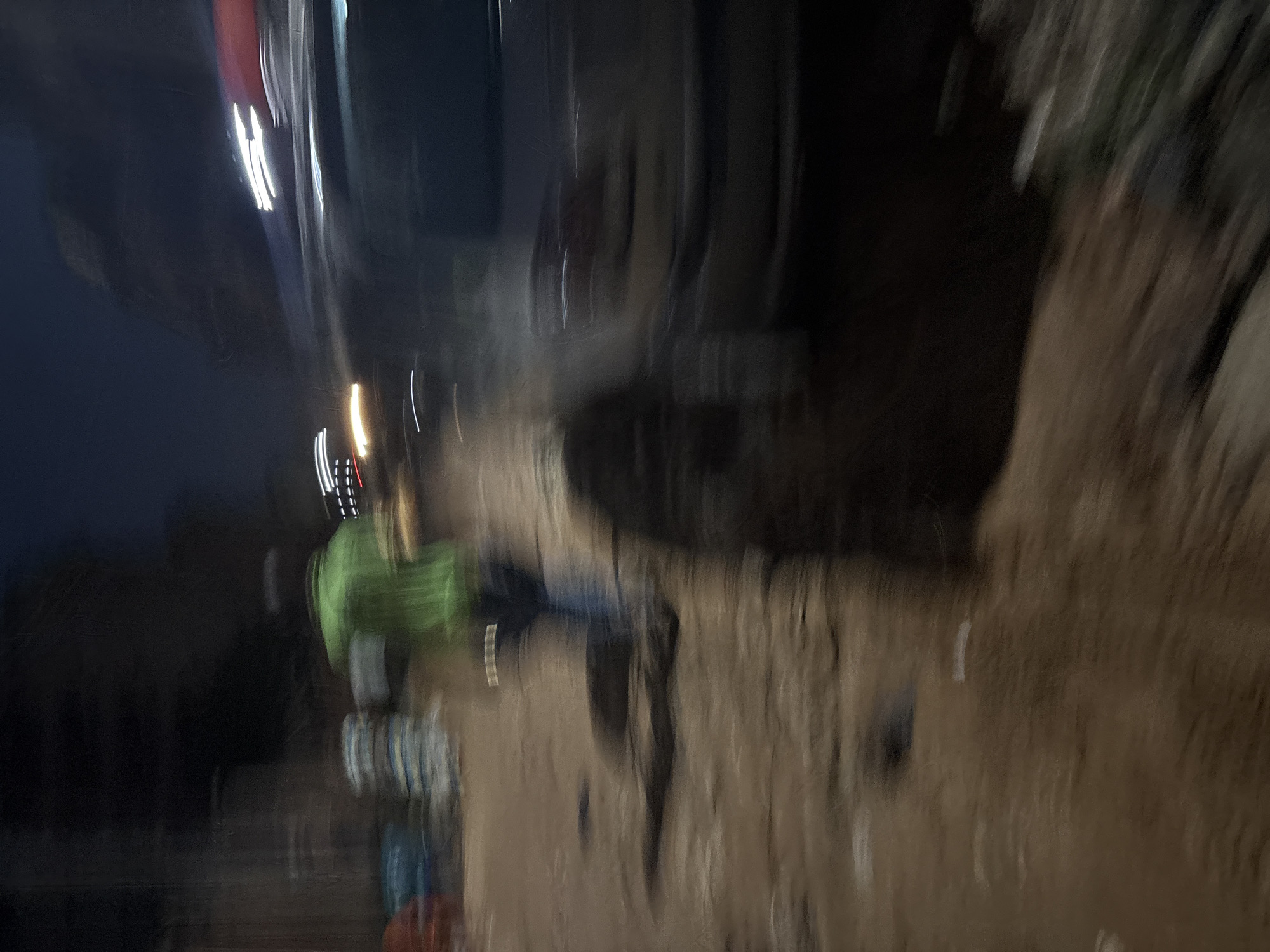 The image depicts a blurred nighttime scene on a dirt road in Onitsha, Nigeria. In the foreground, a person in a bright green shirt and dark pants is captured in motion, their form indistinct due to the blur. To the left, several stacks of what appear to be bagged materials, possibly cement or grain, are visible. A vehicle, also blurred, occupies the right side of the frame, with its headlights and taillights creating streaks of light. The ground is uneven and appears damp, suggesting recent rain or the presence of puddles. The overall impression is one of movement and activity in a dimly lit urban environment.