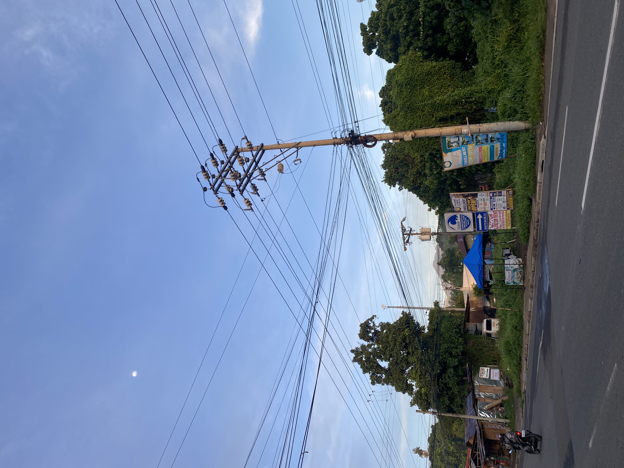 The image captures a street scene in Davao City, Philippines, likely during daytime based on the bright sky and moon visibility. A prominent utility pole with numerous electrical wires dominates the right side of the frame, extending diagonally across the sky. Several trees, lush with green foliage, are visible on either side of the road, with some encroaching on the side of the road and climbing structures.
Along the roadside, several structures appear to be small businesses or temporary stalls. One stall has a blue canopy. Signs are visible, including one with images of people and text that appears to announce enrollment. A white van is parked near these structures. A motorcycle rider is seen on the asphalt road in the foreground, moving from left to right. The road itself shows lane markings. In the distance, mountains can be seen against the clear blue sky, with a faint crescent moon visible. The overall environment suggests a semi-urban or suburban area.