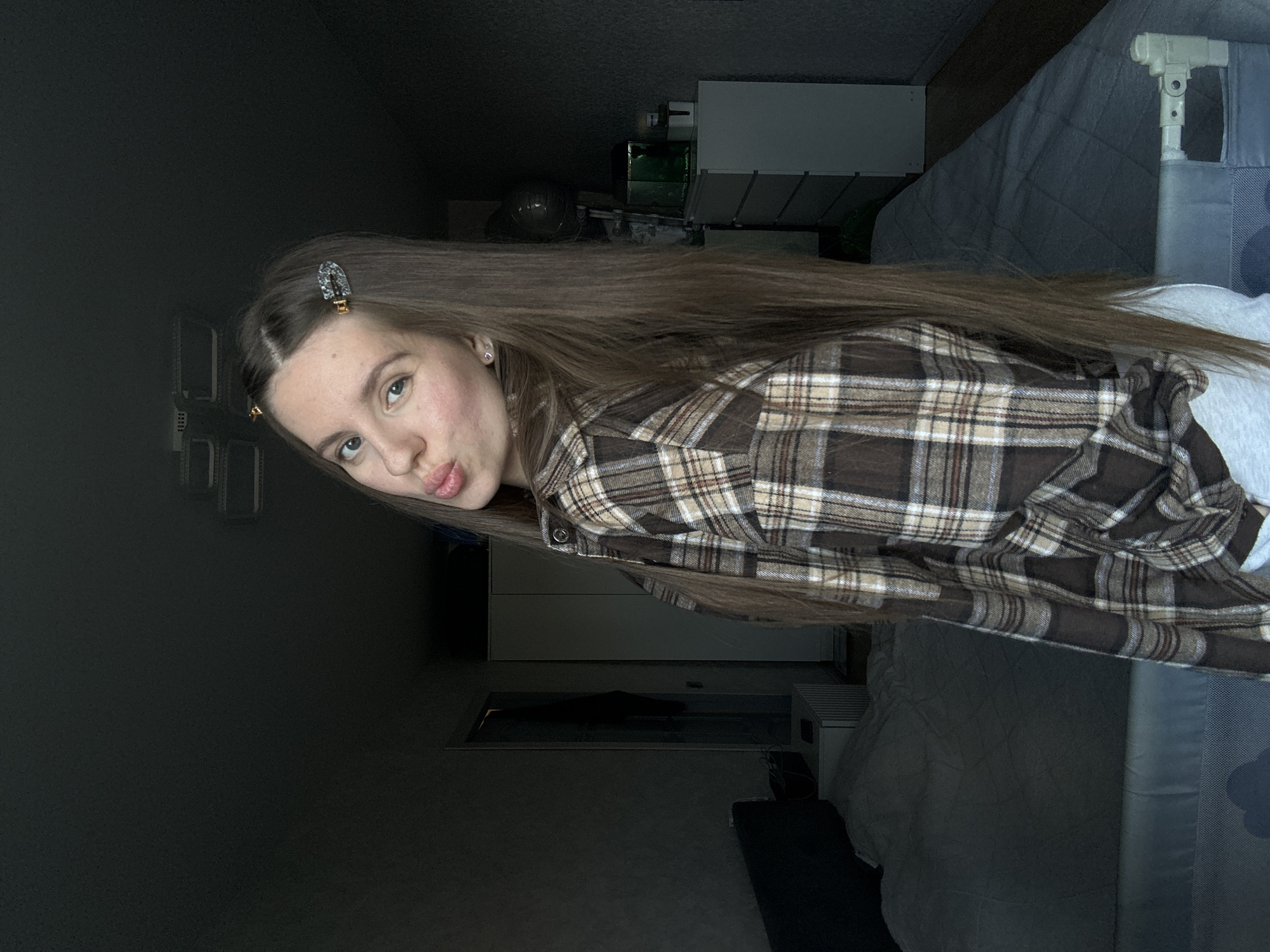 A young woman with long, brown hair and light skin is posing in a dimly lit room. She is wearing a brown and beige plaid flannel shirt and has a decorative hair clip in her hair. The room contains a bed with a grey quilted cover, a white dresser, and various other objects partially visible in the background. The location in the city of Klin, Russia is not determinable from the information present in the image.