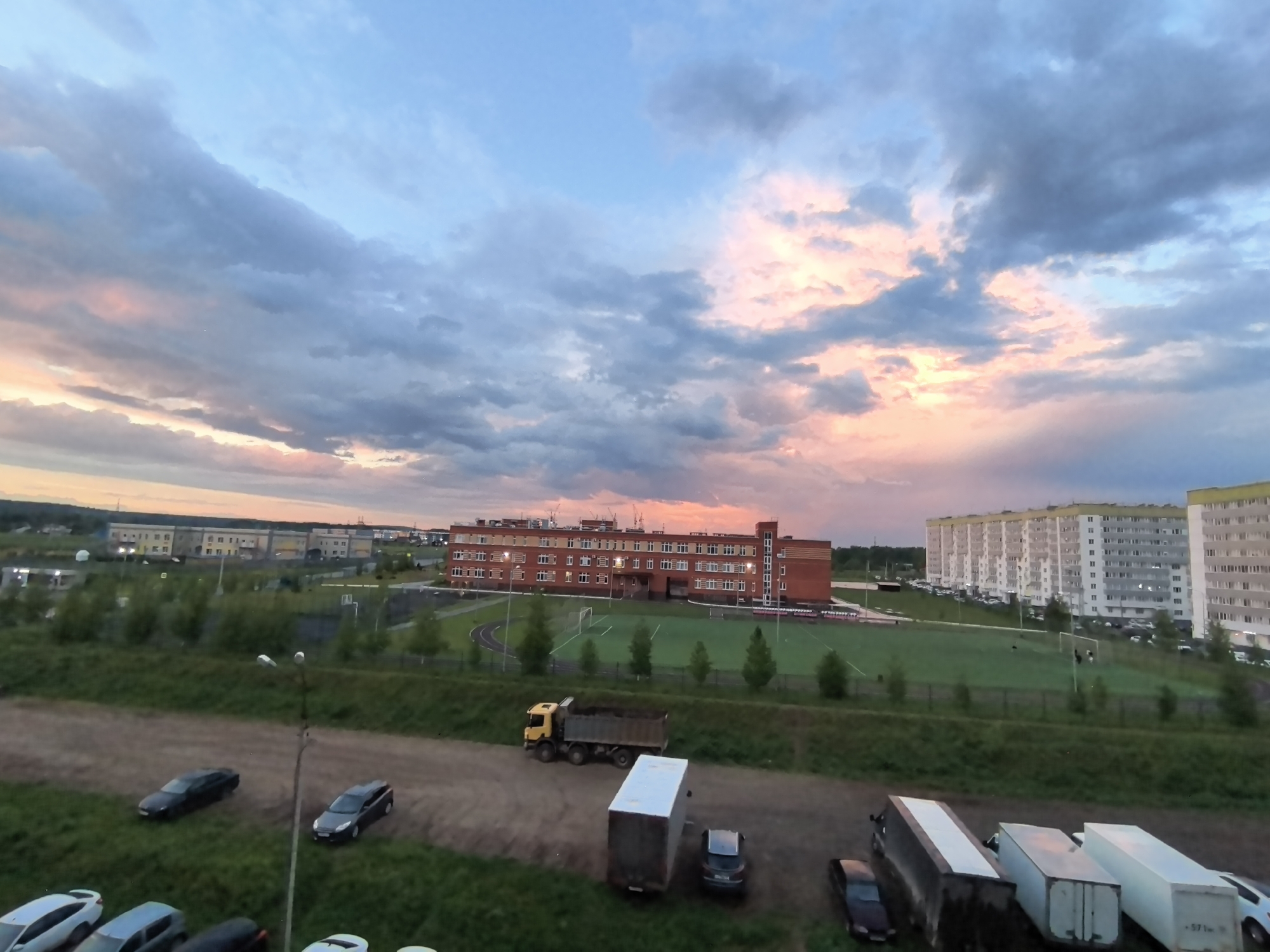 The image presents an elevated view of a scene in Фролы, Russia, under a dramatic sky with pink and gray clouds. In the foreground, a dirt area contains several parked vehicles, including cars and semi-trucks with trailers. A yellow dump truck filled with dirt is also present.
Behind the vehicles, a green area with a line of small trees leads to a red brick building with multiple windows, likely a school or public building. A soccer field with white goalposts and a running track surround the building. To the right of the building are several white apartment buildings. In the far distance, other buildings and structures are visible against the horizon.