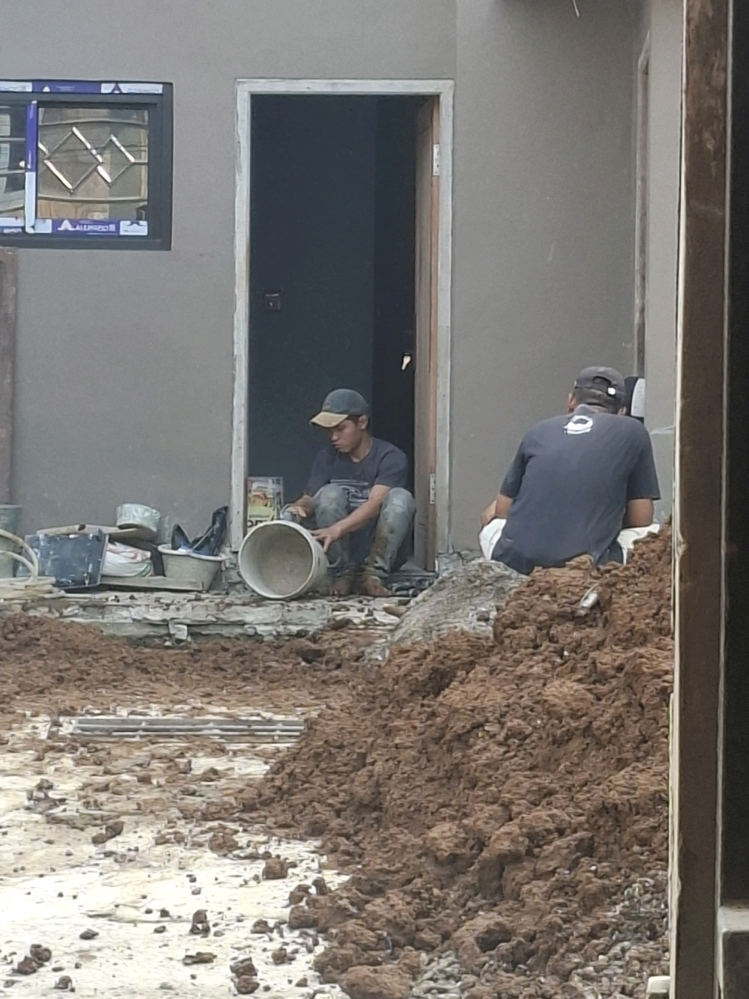 A construction scene in Ciheulang Tonggoh, Indonesia, shows two men working outdoors. The man on the left, wearing a dark t-shirt and a tan cap, is crouched down, tending to a white bucket. In front of him is a collection of construction materials, including buckets, bags, and a metal object. To the right, another man, dressed in a dark t-shirt and a dark cap, is seated on a pile of dirt, facing away from the camera. He is wearing white pants. The background reveals the partially constructed exterior of a building with a dark doorway and a window. The ground is covered in dirt, debris, and broken bricks, indicating ongoing construction work.